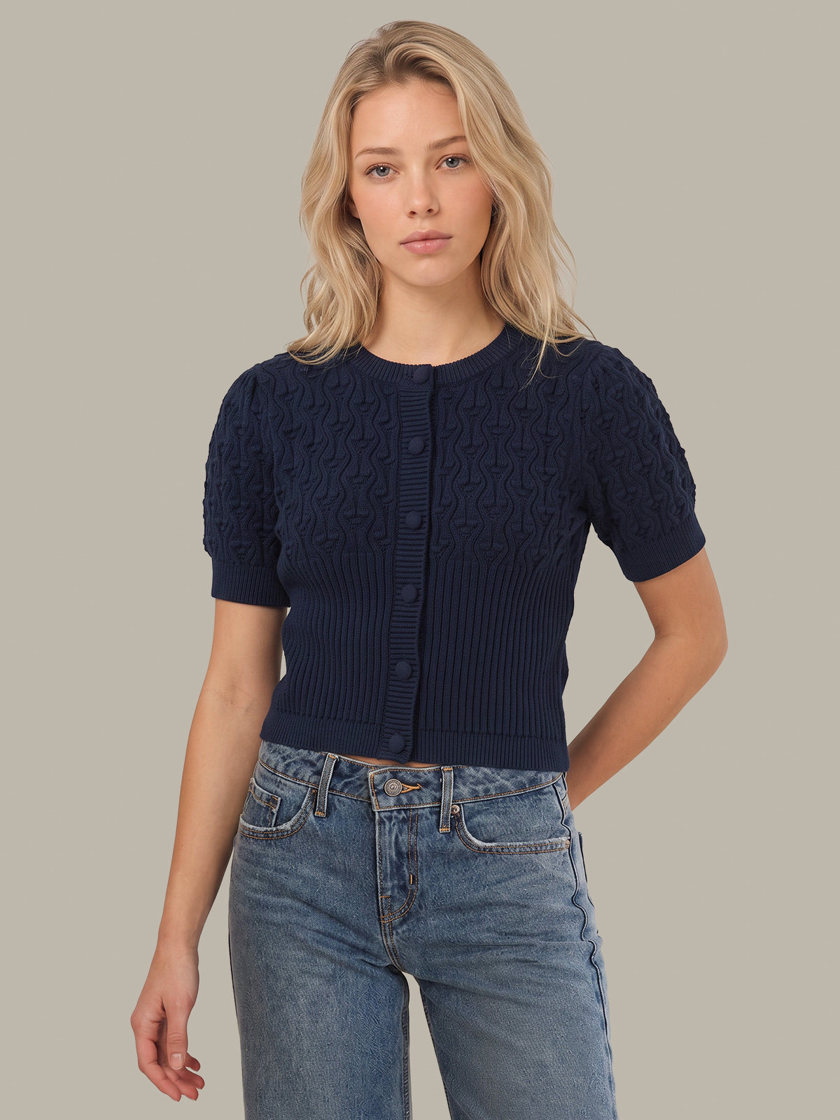 A blonde woman wearing a navy blue short-sleeve cable-knit cardigan with button-front closure and blue jeans, standing against a solid light gray studio background.