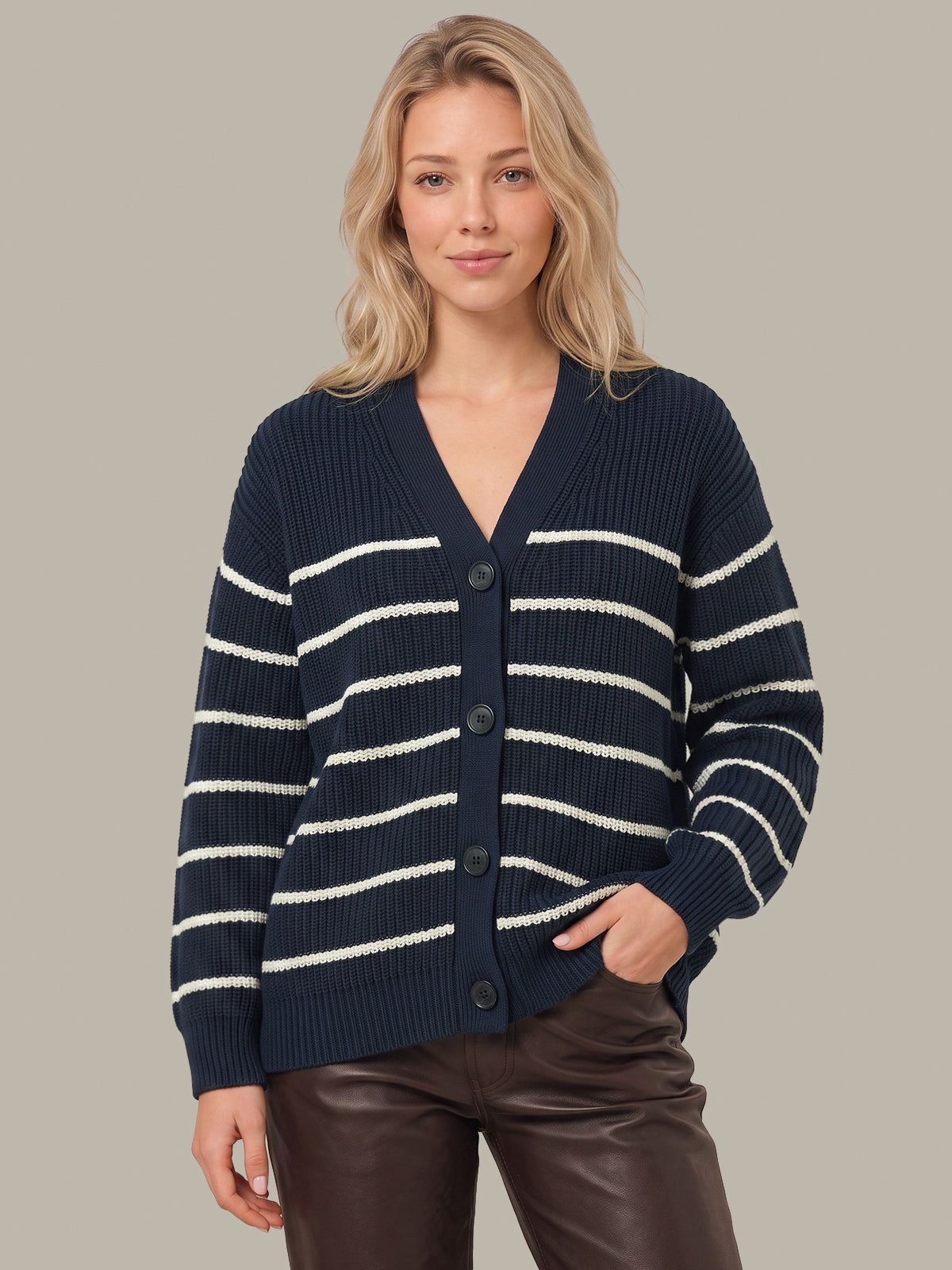 A woman wearing a navy blue ribbed cardigan with horizontal cream stripes and dark brown faux-leather pants, standing against a solid light gray background.