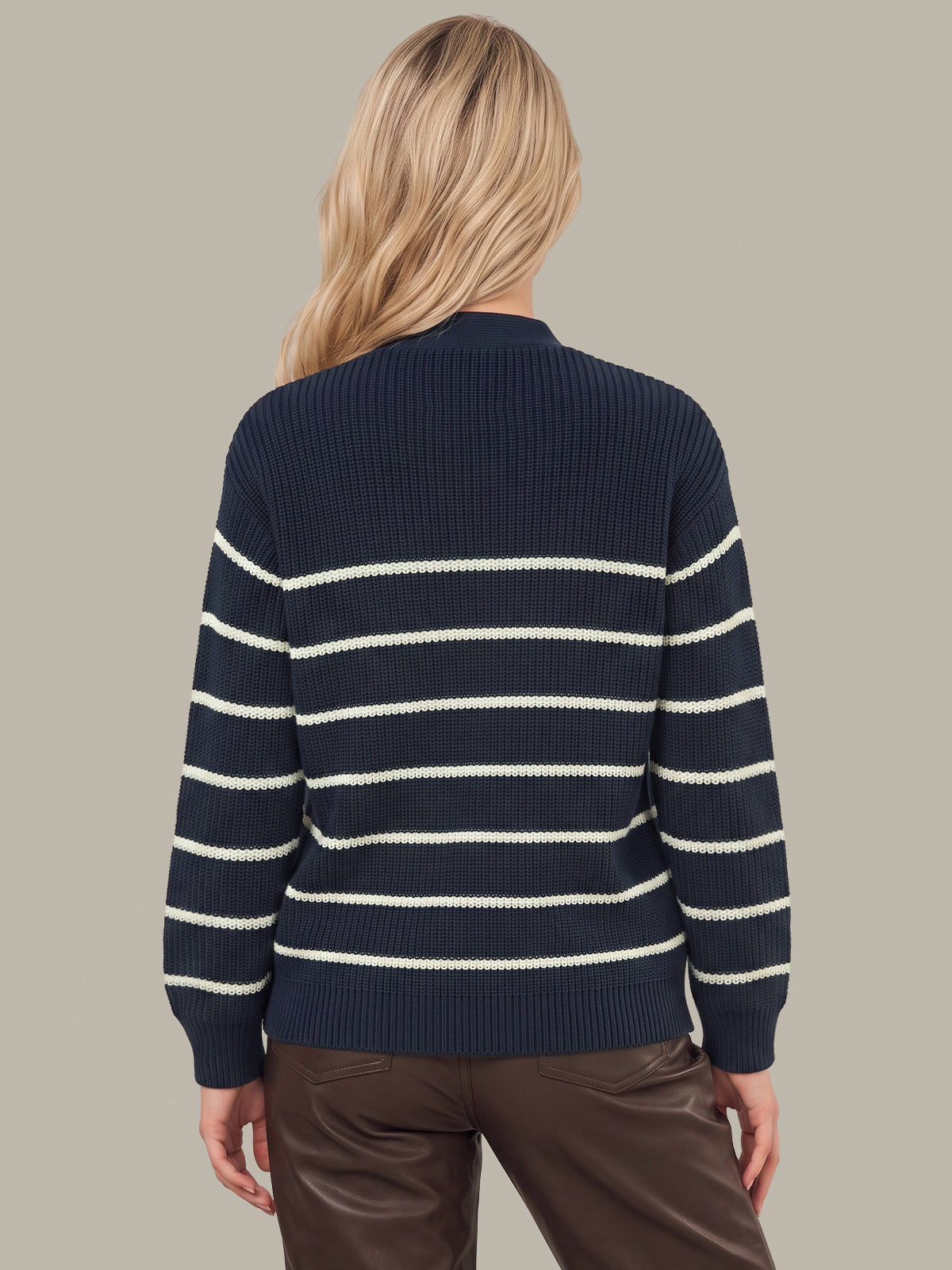 A woman viewed from the back wearing a navy blue ribbed knit sweater with horizontal cream stripes and brown pants, set against a solid taupe background.