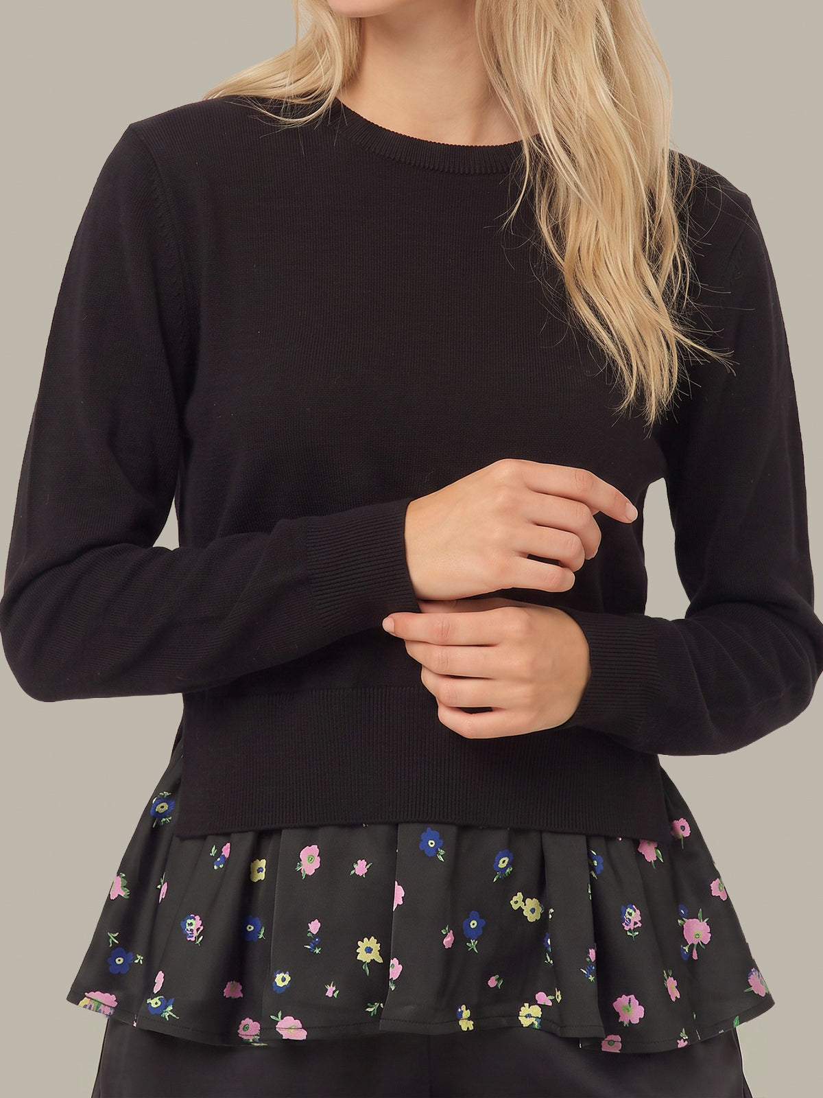 A woman wearing a black long-sleeve knit top with a black peplum hem featuring a small multicolor floral print, shown against a plain light gray background.