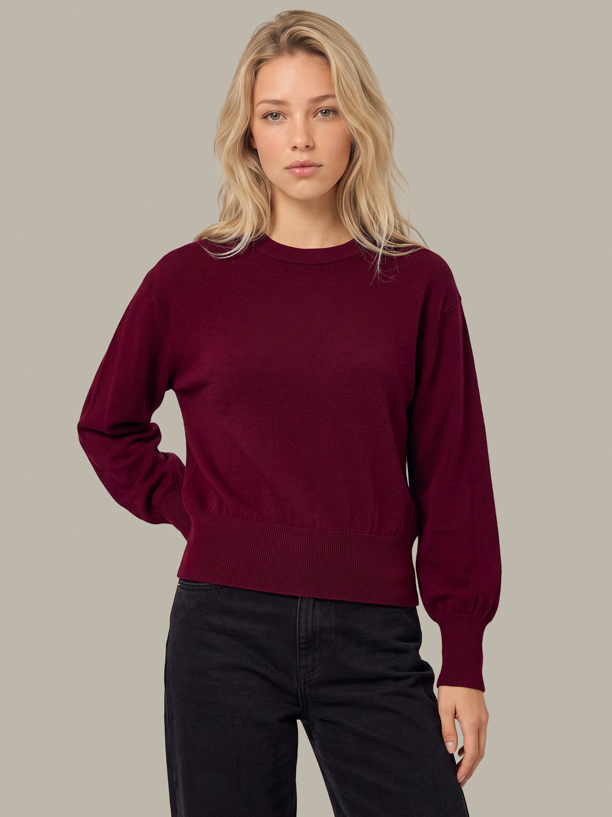 A blonde woman wearing a dark burgundy long-sleeve crewneck sweater and black jeans, standing against a solid light gray background.