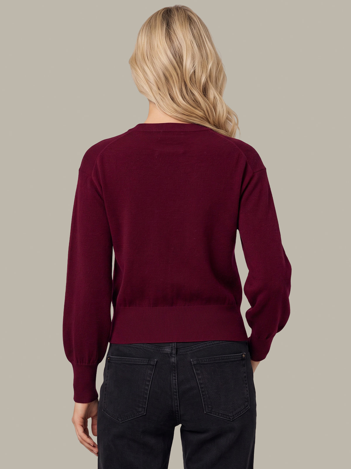 A woman viewed from the back wearing a long-sleeve crewneck sweater in deep burgundy and dark jeans, set against a solid light gray background.