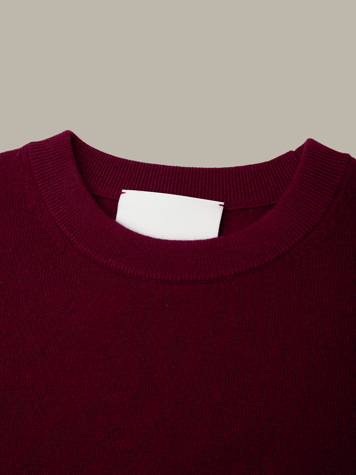 Close-up of a crewneck sweater in deep burgundy, showing the ribbed collar and a blank white neck label against a solid taupe background.