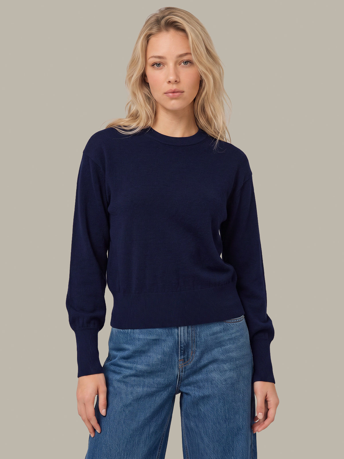 A blonde woman wearing a dark navy blue long-sleeve crewneck sweater and blue jeans, standing against a solid light gray background.
