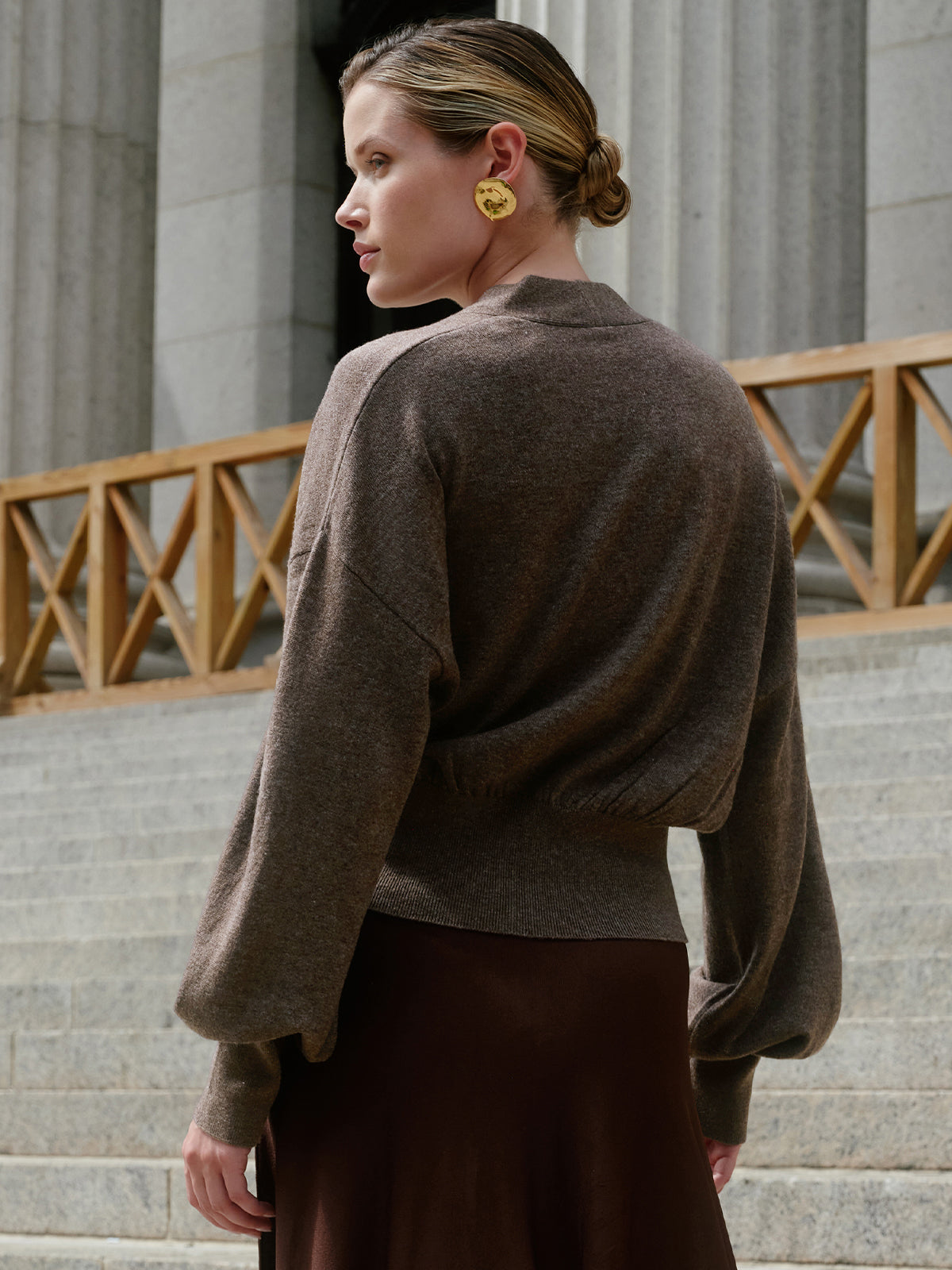 A woman wearing a brown knit sweater and a dark brown skirt, photographed from the back in an outdoor setting with stone steps and columns.