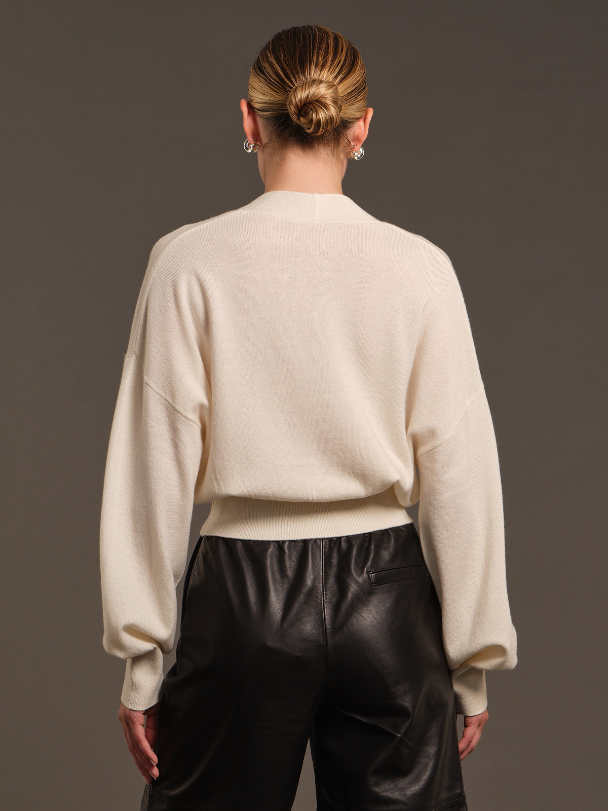 A model shown from the back wearing a cream-colored knit sweater and black leather pants against a solid dark gray studio background.