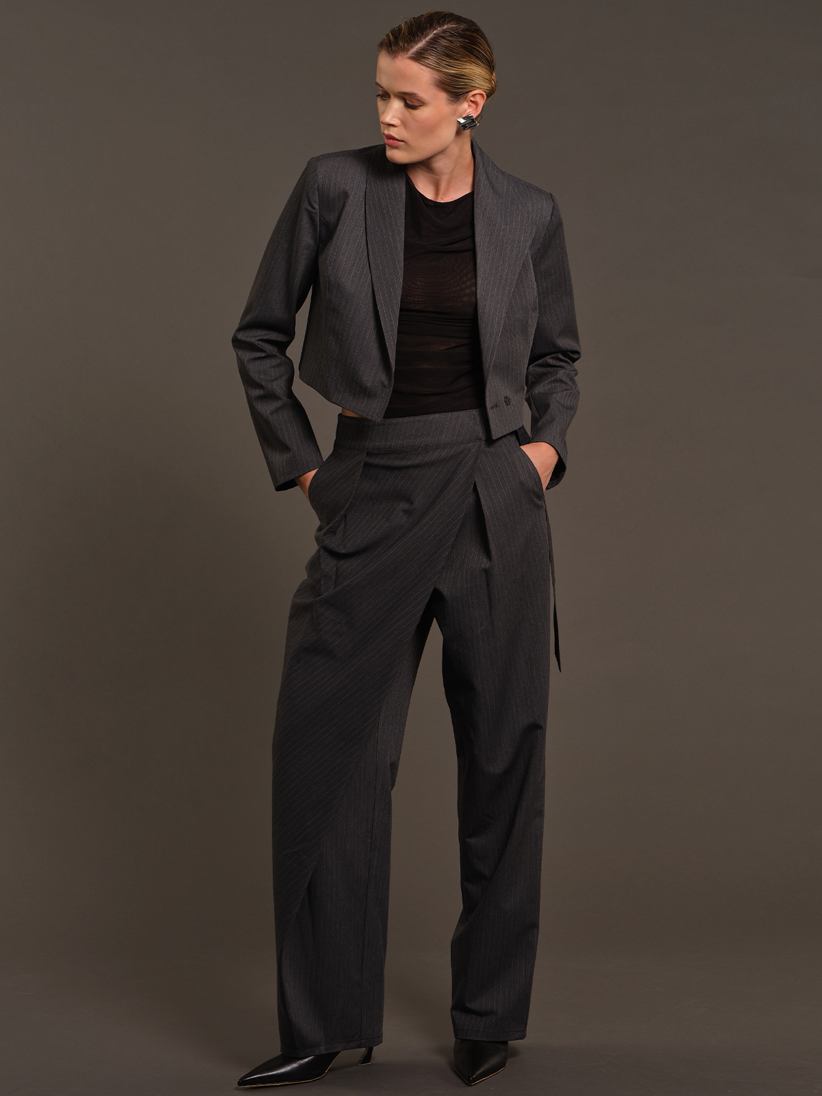 A model wearing a dark gray pinstripe cropped blazer, matching wide-leg trousers, and a black top, standing against a solid dark gray studio background.