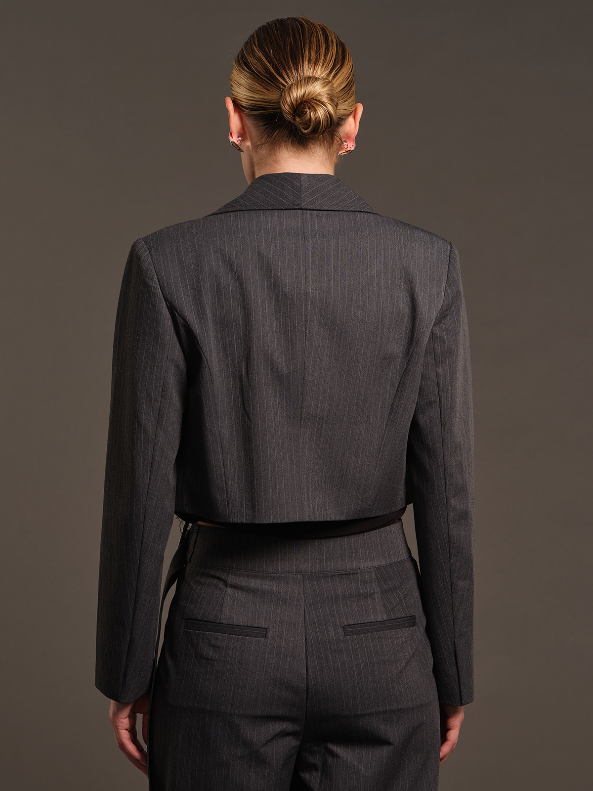 A model shown from the back wearing a dark gray pinstripe cropped blazer and matching trousers against a solid dark gray studio background.