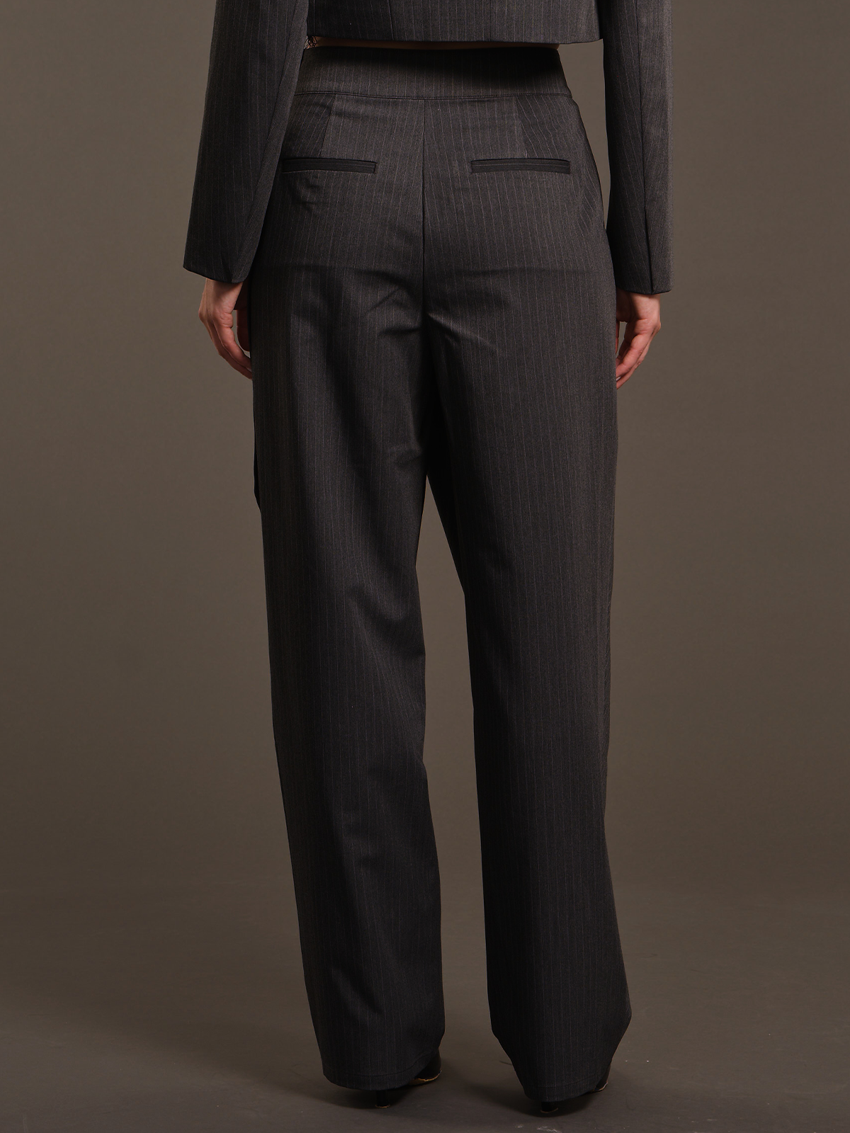 Rear view of a model wearing dark gray pinstripe trousers and a matching dark gray cropped blazer against a solid taupe background.