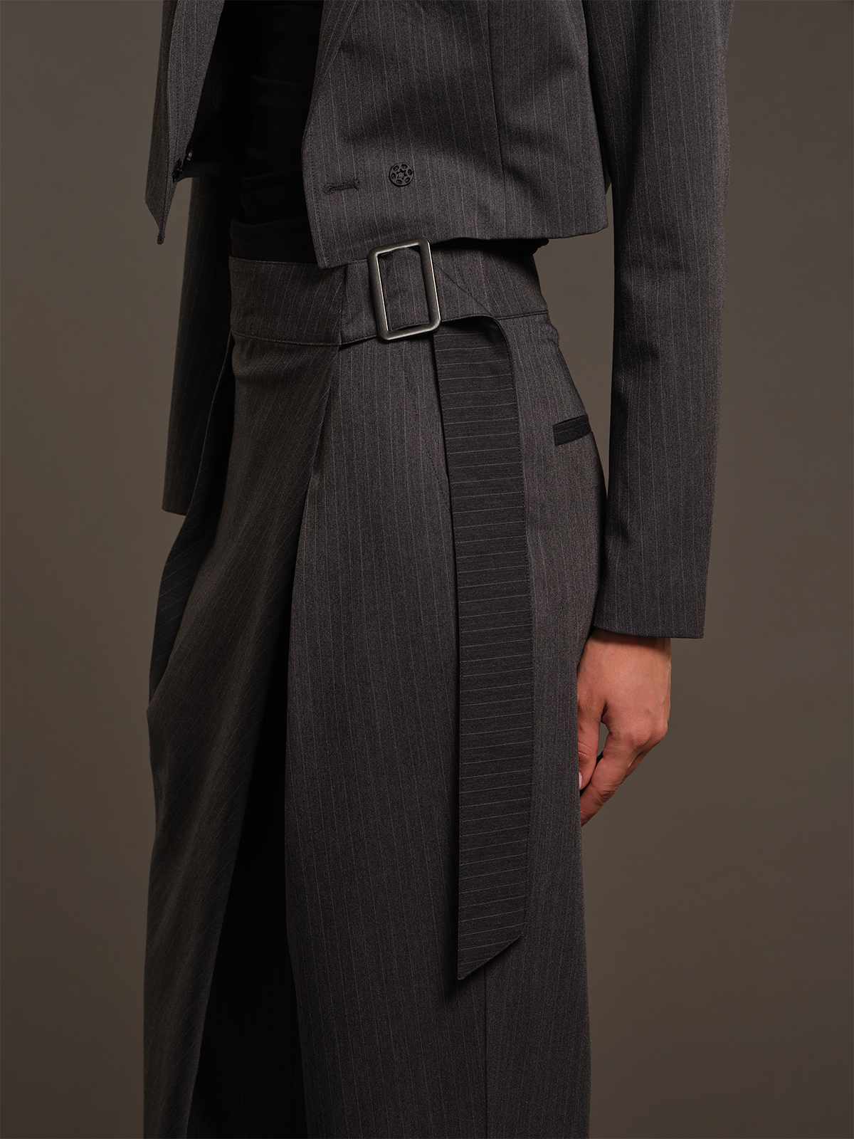A close-up of a model wearing a dark gray pinstripe suit, featuring a cropped blazer with a small embroidered logo on the chest, a matching high-waisted wrap skirt with a belt and silver buckle, and a black top underneath.