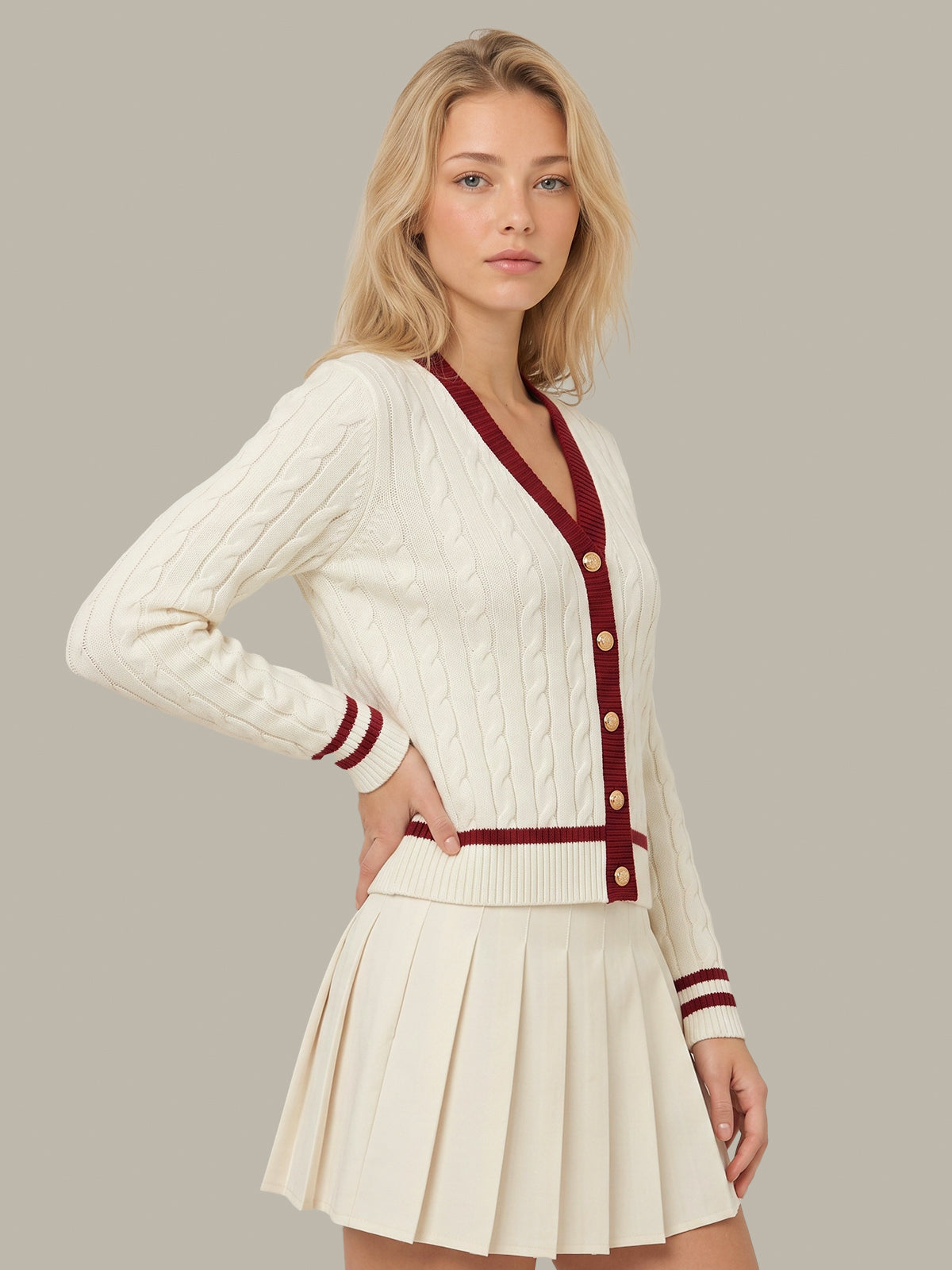 A blonde woman models a cream-colored cable-knit cardigan with burgundy trim and buttons, paired with a matching pleated mini skirt, against a solid gray studio background.