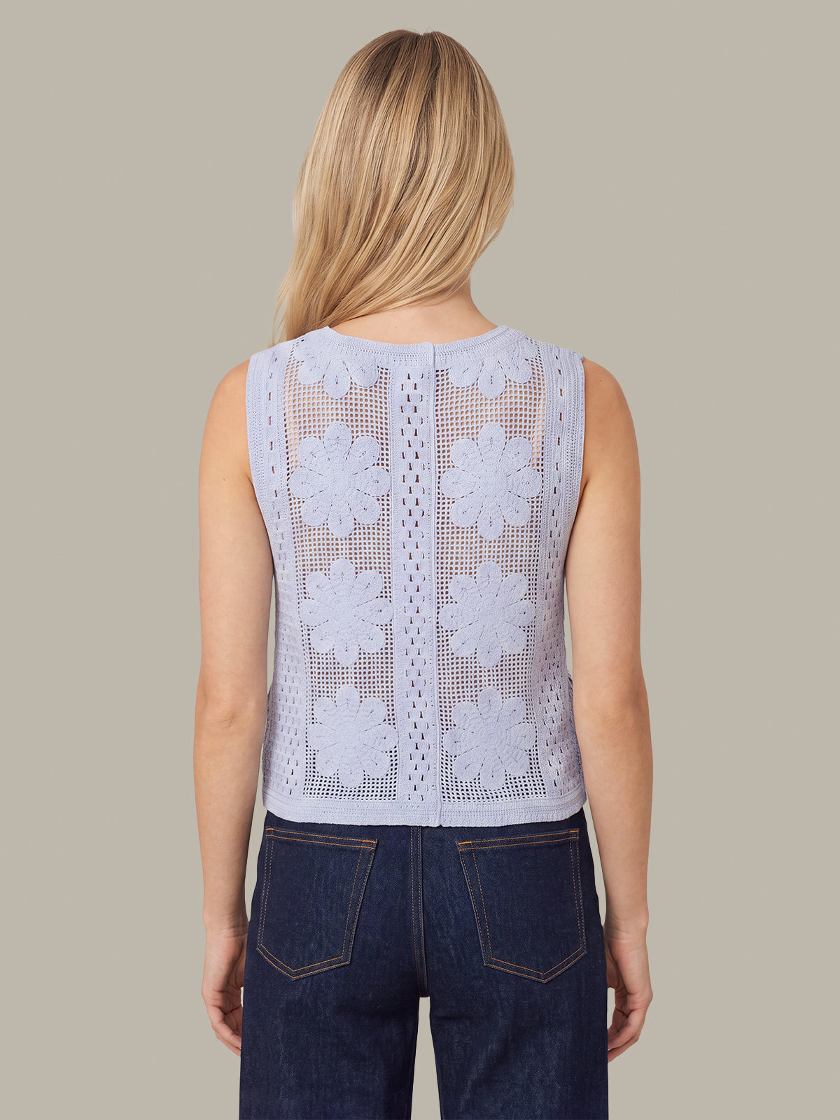 A woman viewed from the back wearing a sleeveless white crochet lace top with floral patterns and dark blue jeans, set against a solid taupe background.