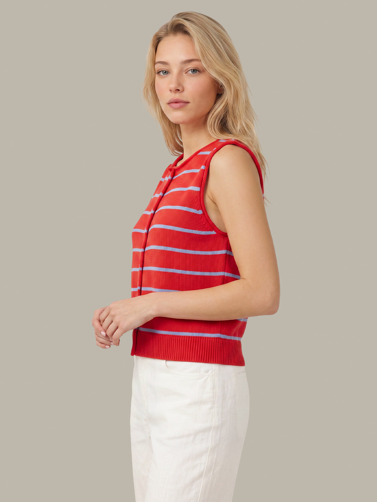 A blonde woman wearing a red sleeveless knit top with thin light blue horizontal stripes and white pants, standing against a solid light gray studio background.