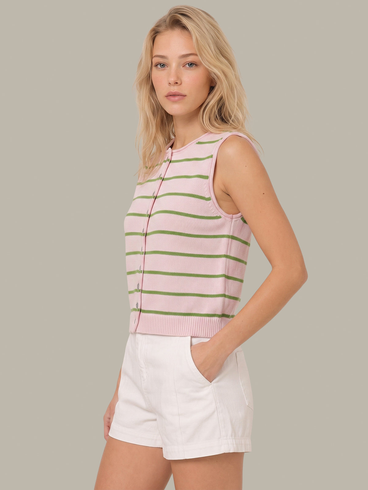 A woman modeling a sleeveless knit top with horizontal pink and green stripes, paired with white shorts, against a solid light gray background.