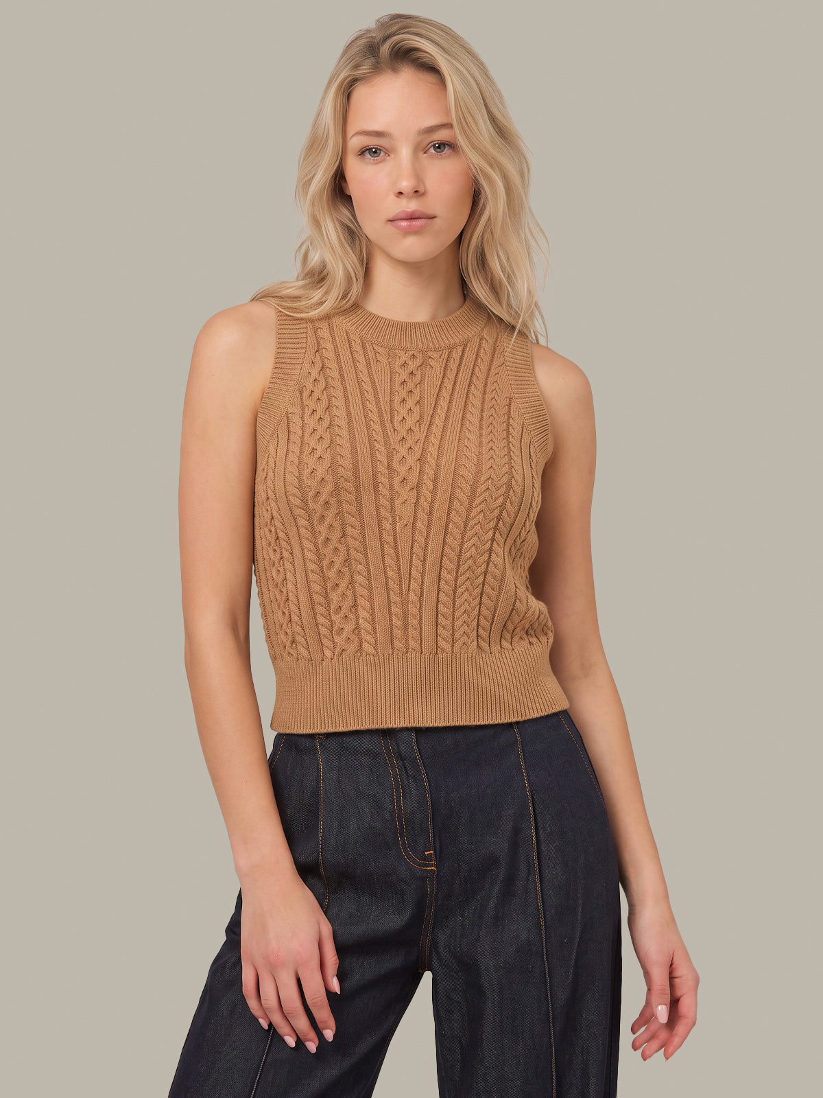 A woman modeling a sleeveless ribbed knit sweater in camel tan, paired with dark blue jeans, against a solid light gray background.