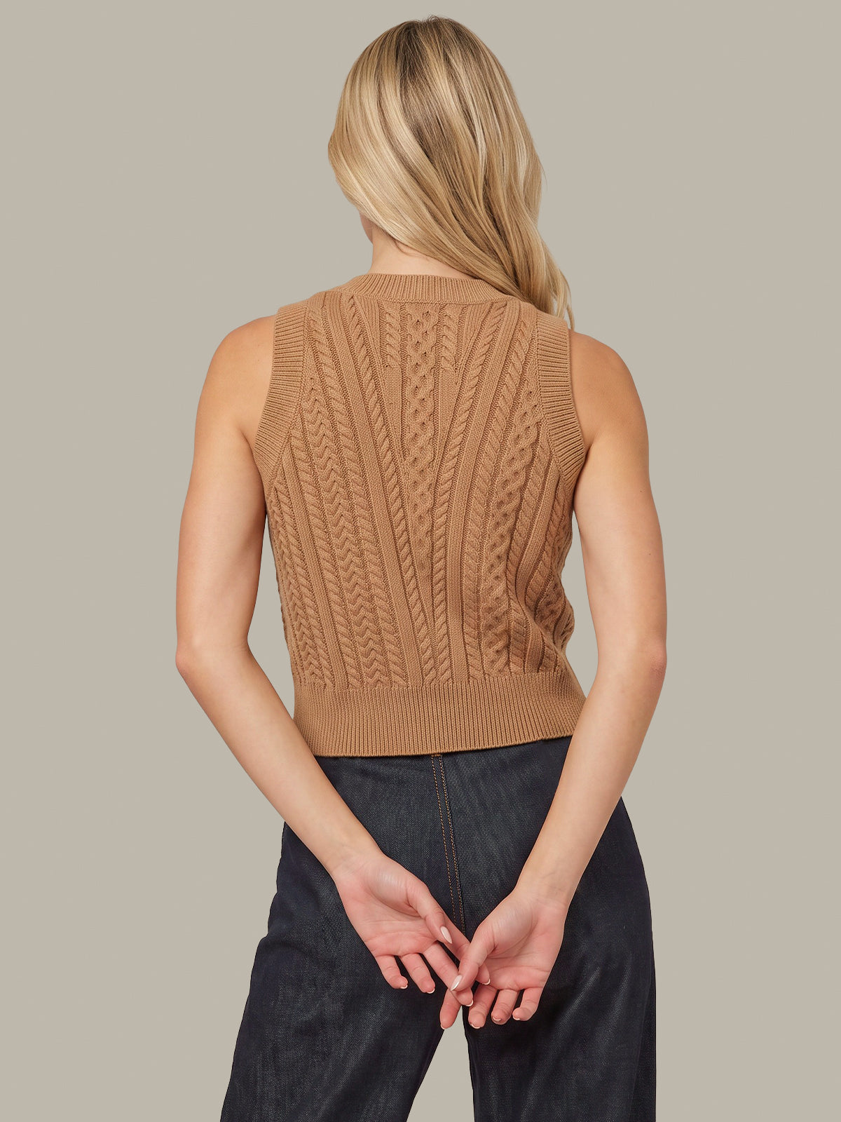 A woman shown from the back wearing a sleeveless ribbed knit sweater vest in camel brown, paired with dark jeans, against a solid light gray background.