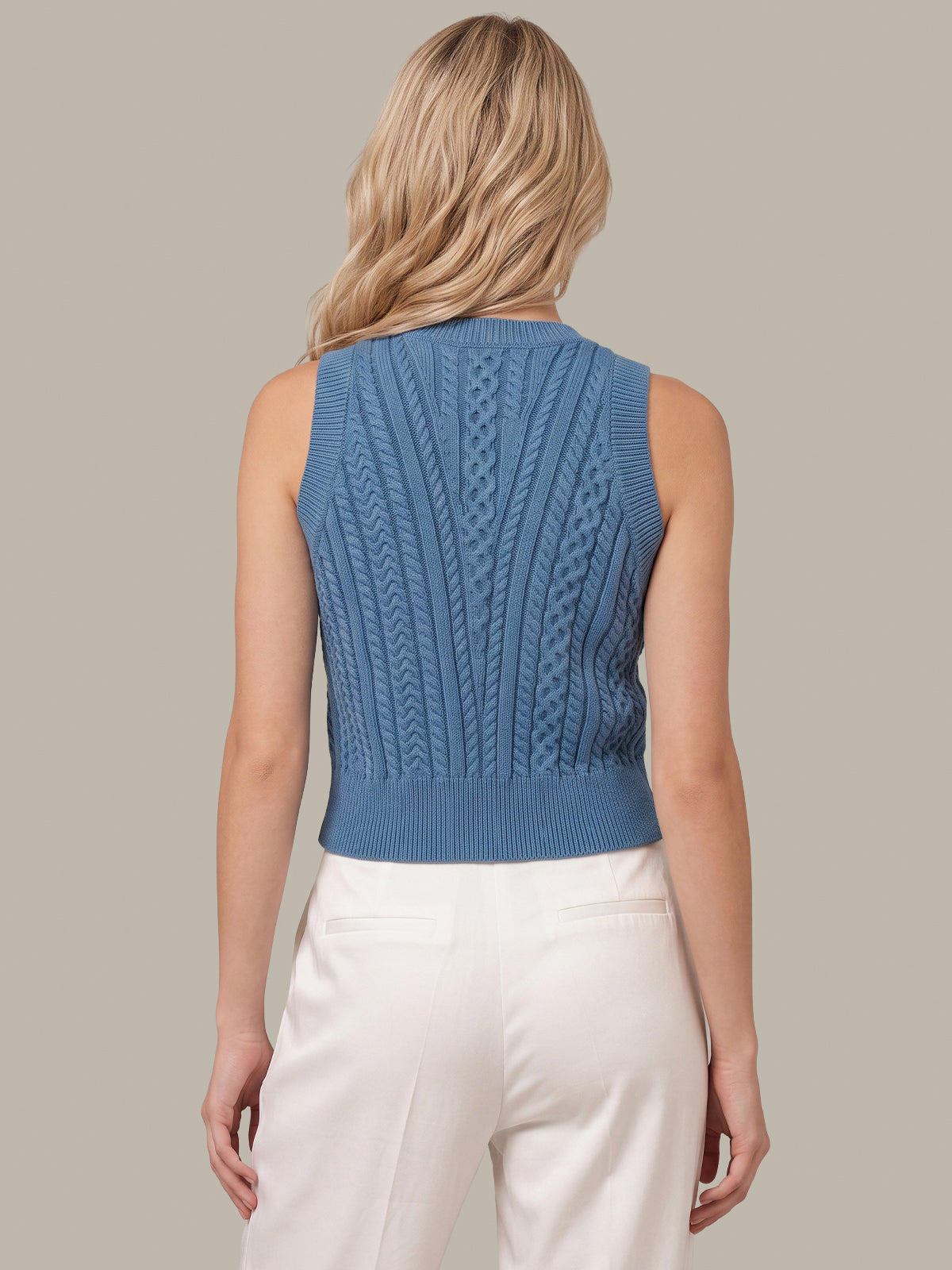 A woman shown from the back wearing a sleeveless blue cable-knit cropped sweater vest and white pants, against a solid light gray background.