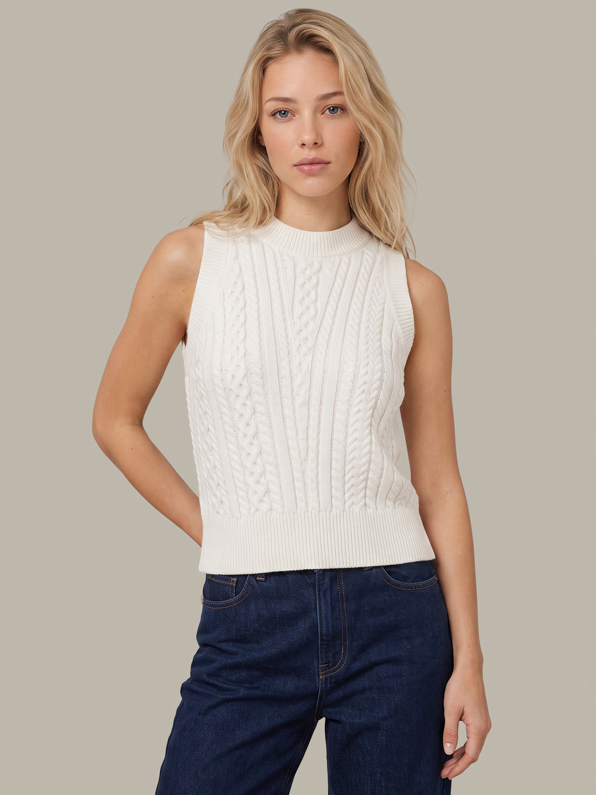A blonde woman wearing a sleeveless white cable-knit mock neck sweater vest paired with dark blue jeans, standing against a solid light gray background.
