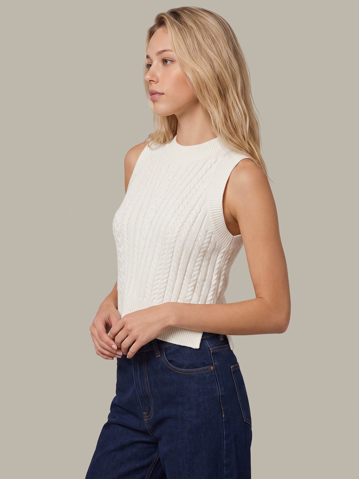 A blonde woman wearing a sleeveless white cable-knit mock-neck top and dark blue jeans, standing against a solid light gray background.