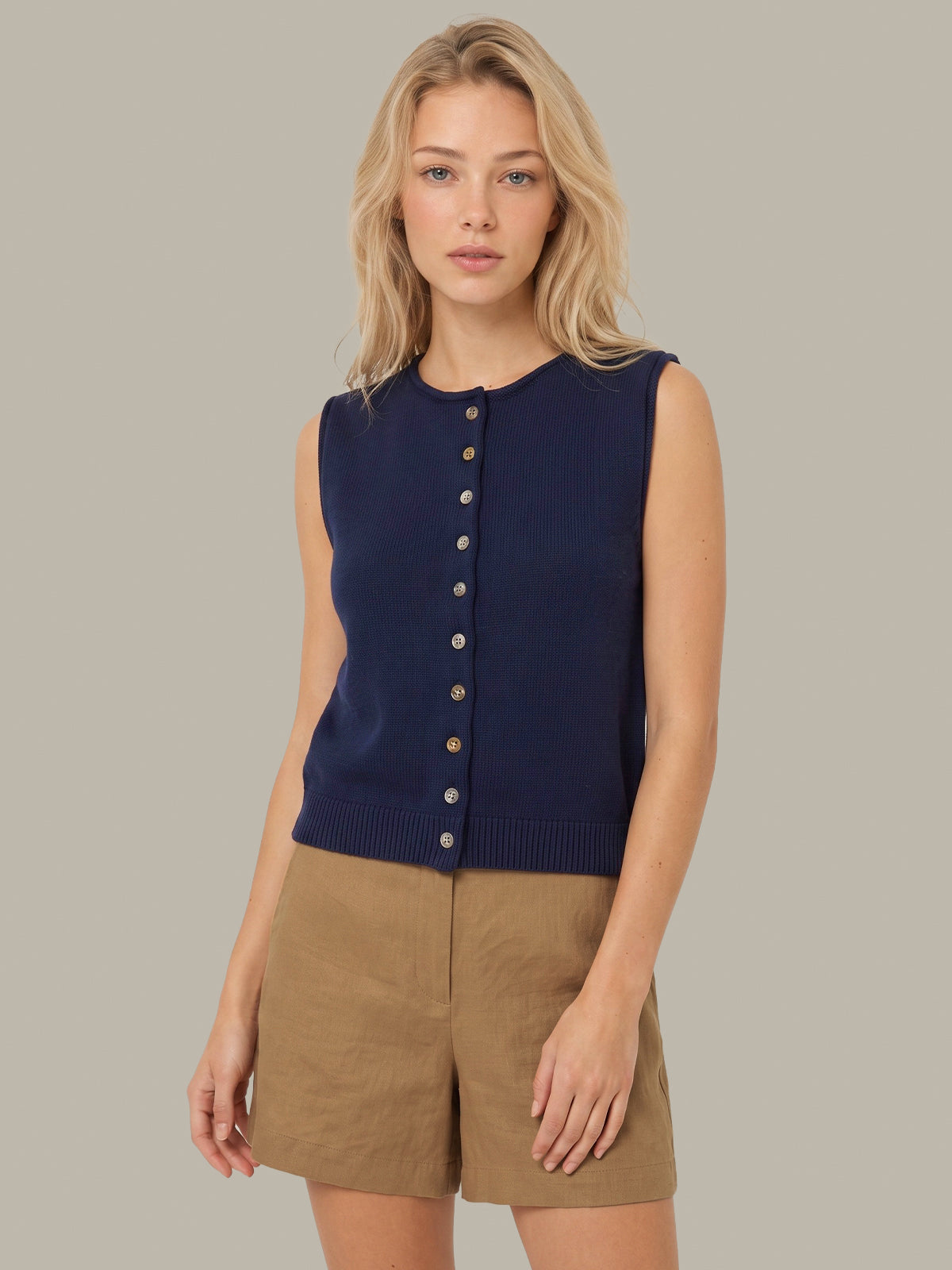 A blonde woman modeling a sleeveless navy blue knit button-front vest with tan shorts, set against a solid neutral gray background.