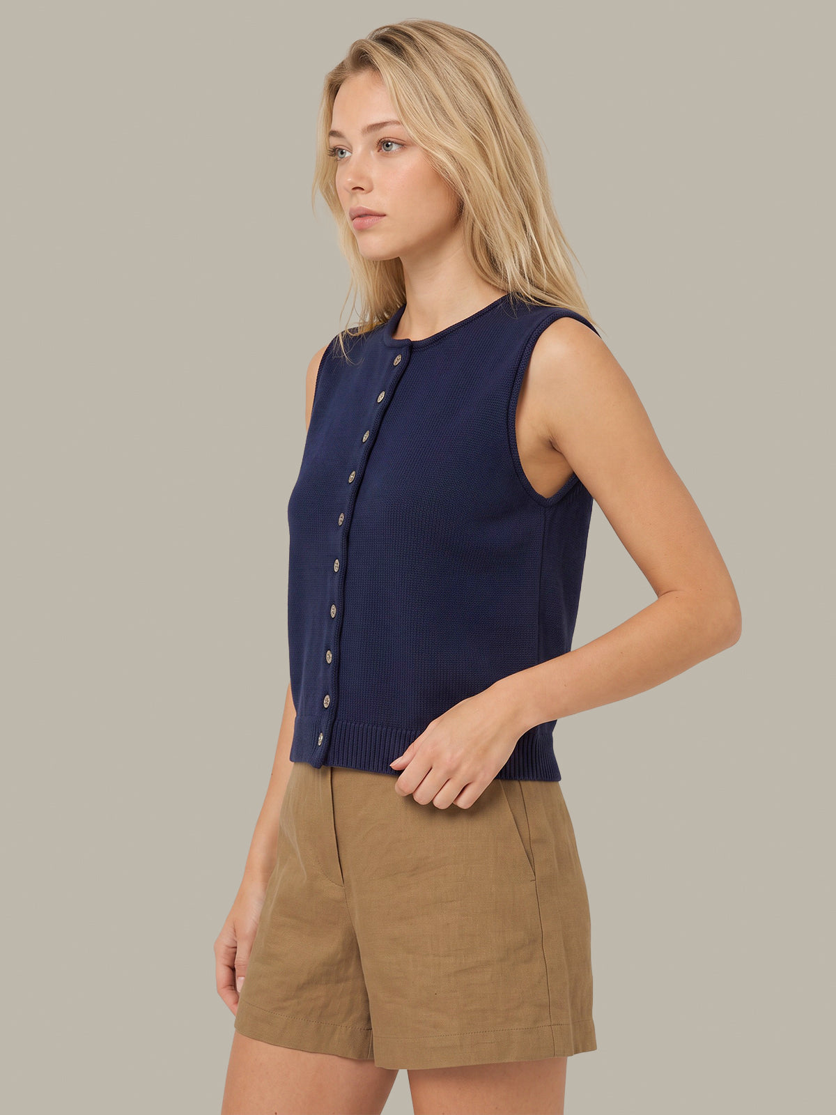A woman modeling a sleeveless navy blue knit top with a button front and tan shorts against a solid gray studio background.