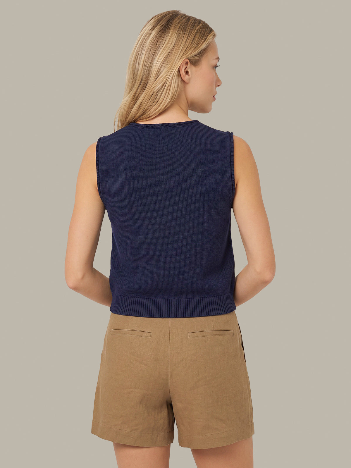 A woman shown from the back wearing a navy blue sleeveless knit top and tan shorts against a solid light gray background.