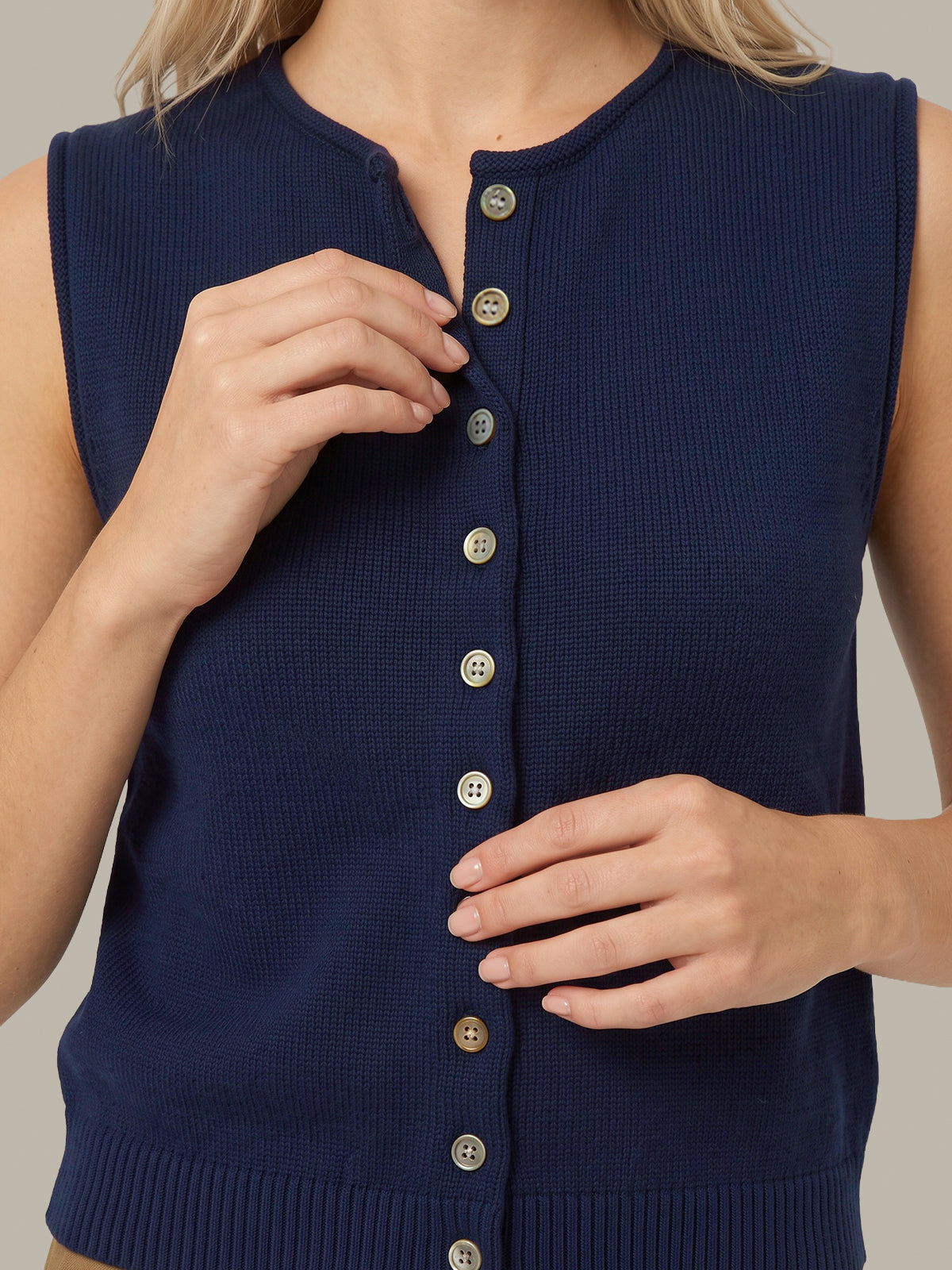 A close-up of a woman wearing a sleeveless navy blue ribbed knit vest with a button-front closure. Her hands are adjusting the top button, and the background is a solid light gray.