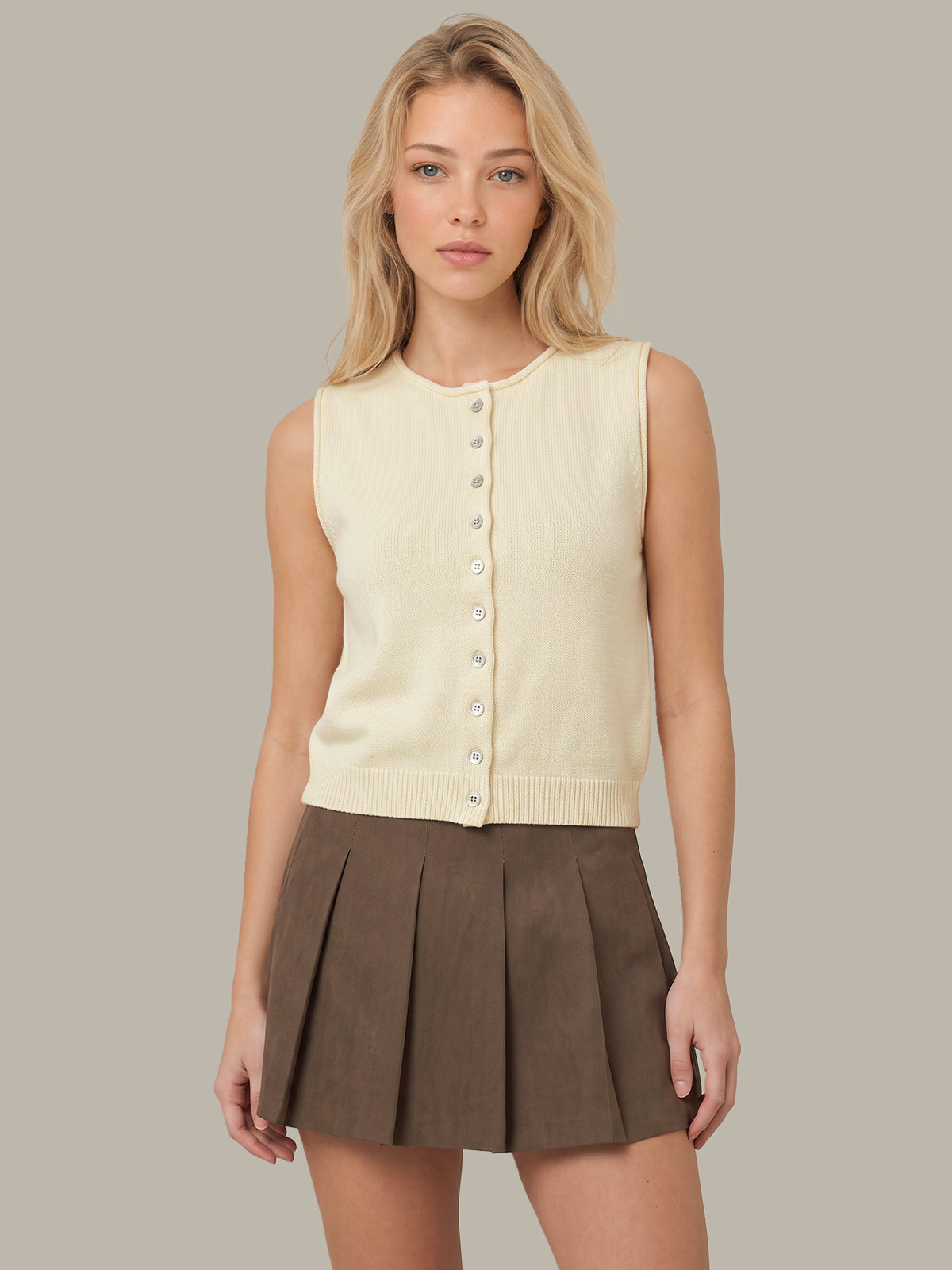 A woman modeling a cream sleeveless button-front knit top paired with a brown pleated mini skirt, set against a solid light gray studio background.