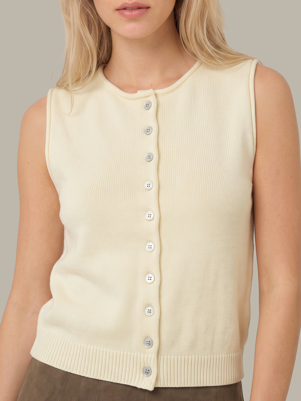 A woman wearing a sleeveless cream ribbed knit vest with a front button closure, shown from the shoulders to the waist against a plain light gray background.