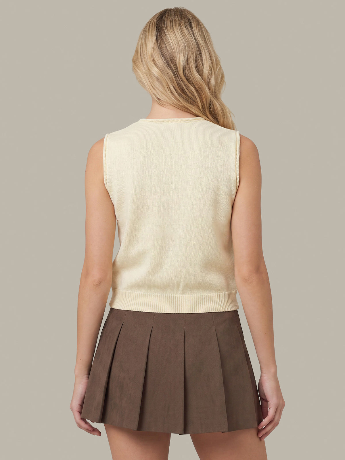 A woman shown from the back wearing a cream sleeveless knit sweater vest and a brown pleated mini skirt, set against a solid taupe background.