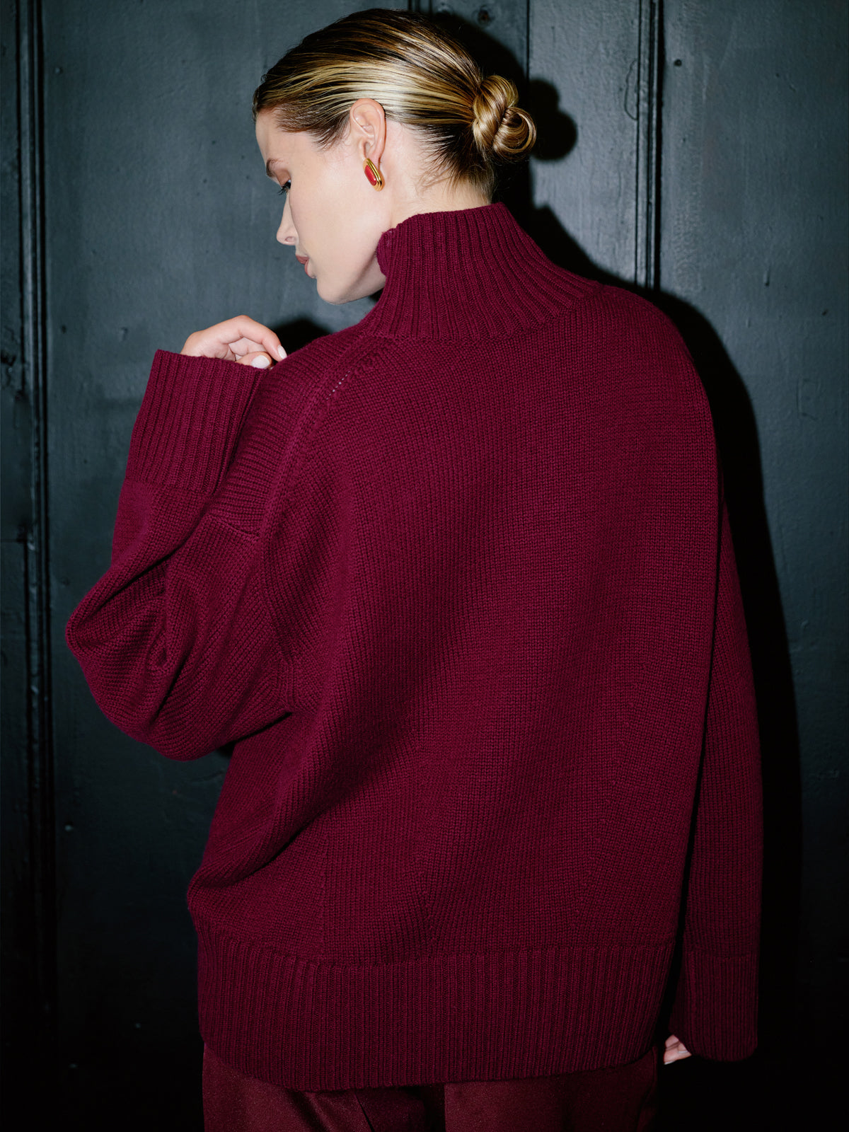 A woman wearing an oversized ribbed turtleneck sweater in deep burgundy, shown from the back against a dark textured wooden wall.