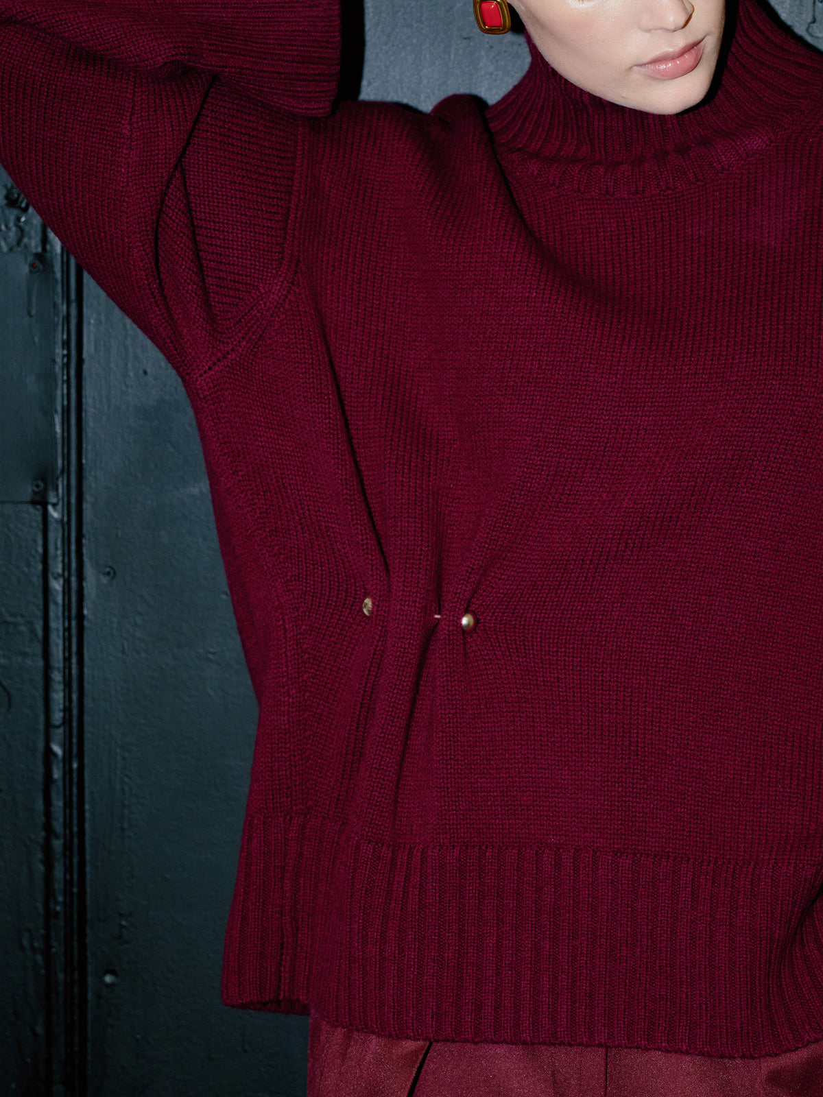 A close-up of a person wearing a ribbed knit turtleneck sweater in deep burgundy, with a dark textured wall behind them.