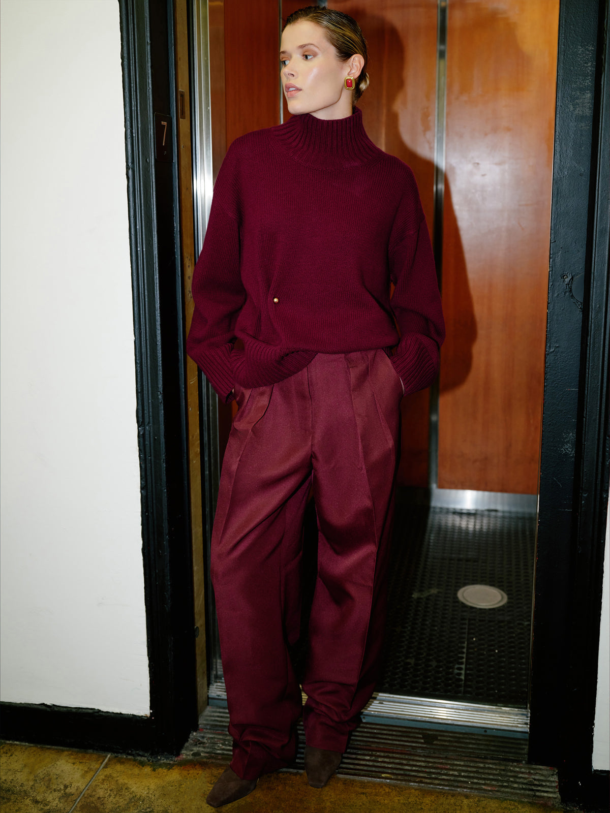 A model wearing a matching burgundy knit turtleneck sweater and wide-leg trousers, standing in a doorway.