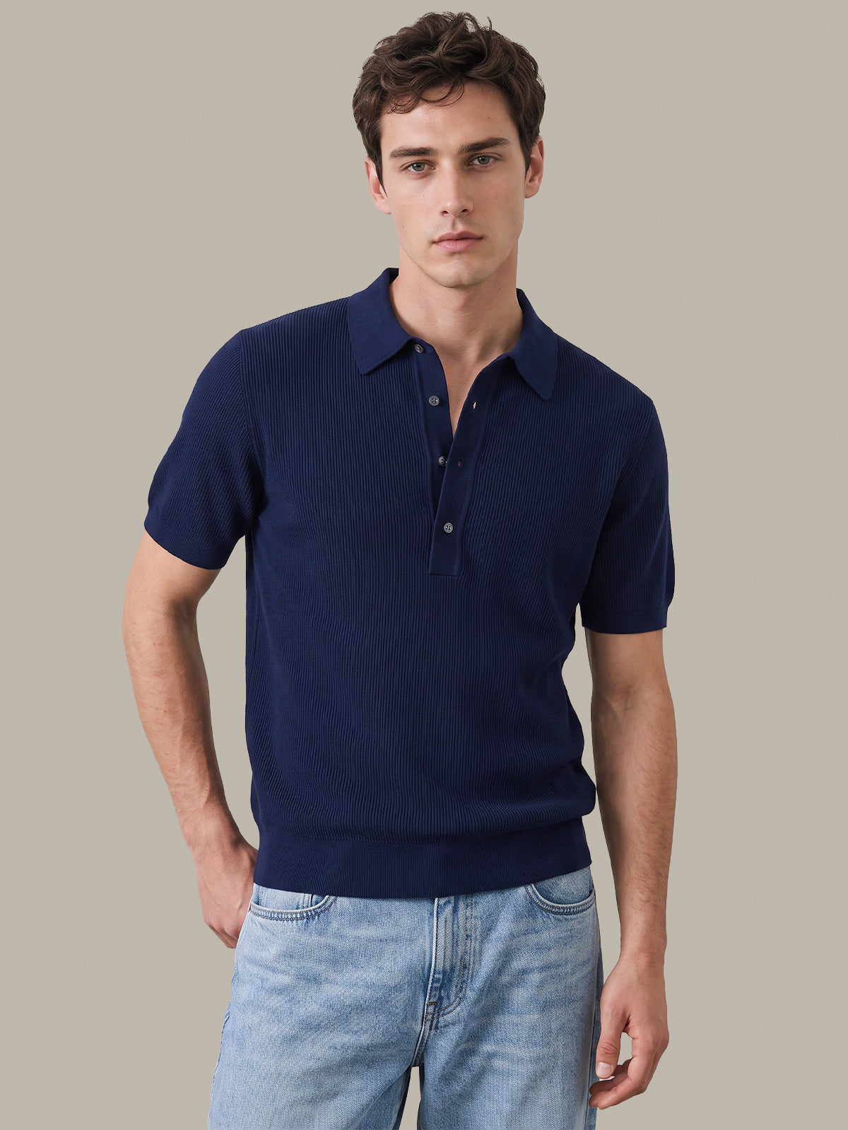A man wearing a dark navy blue ribbed short-sleeve polo shirt and light blue jeans, standing against a solid light gray background.