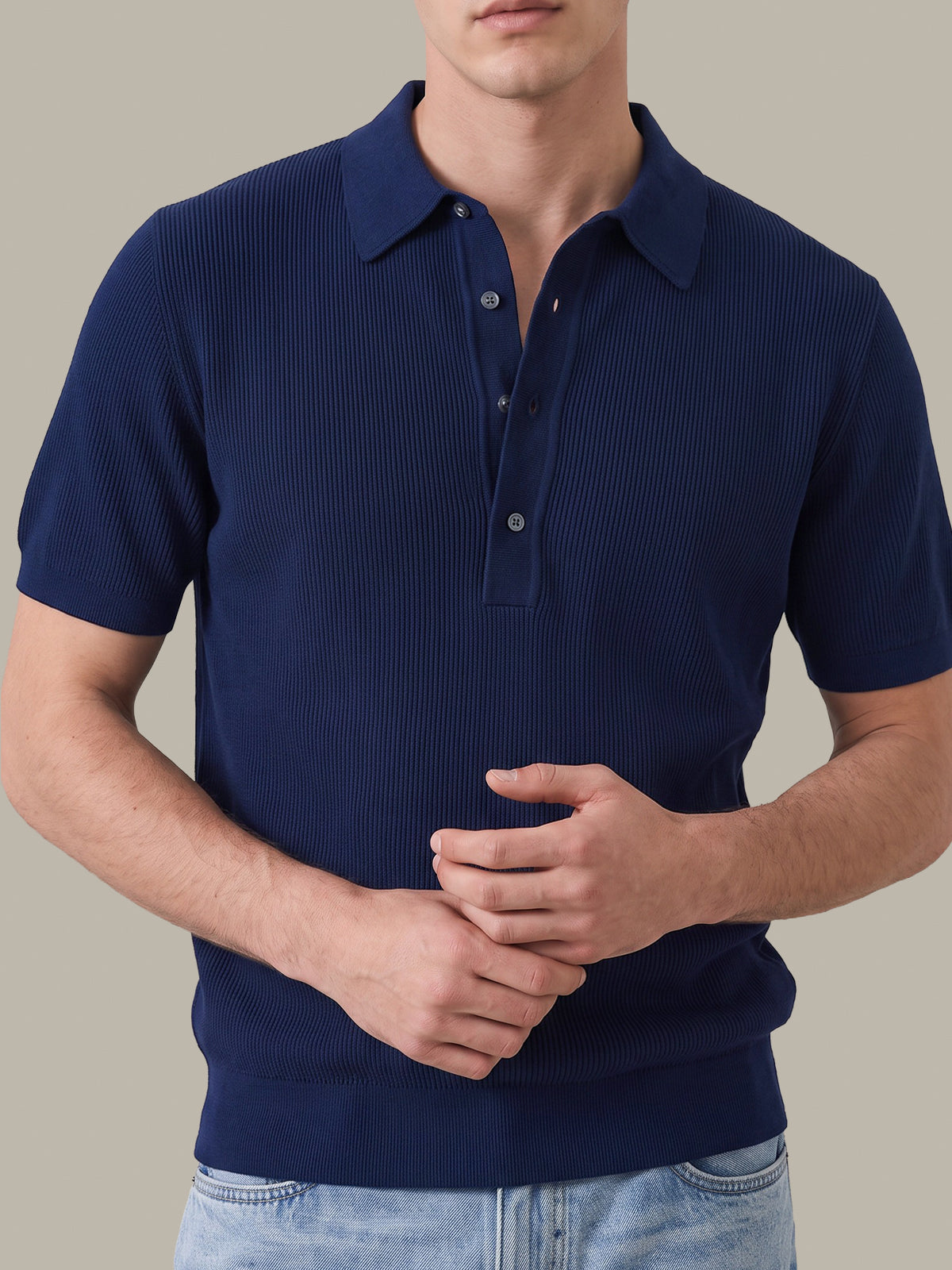 A man wearing a navy blue short-sleeve ribbed knit polo shirt with a three-button placket, paired with light blue jeans, against a solid beige background.