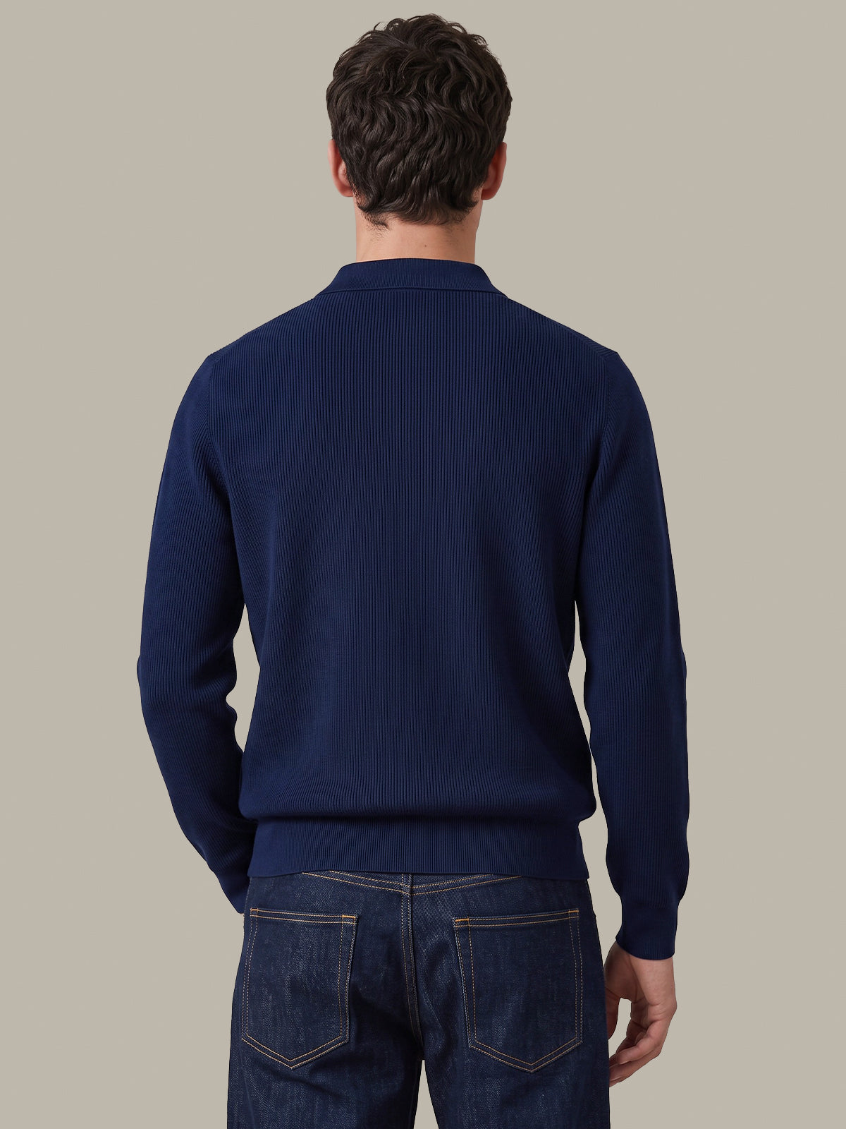 A man viewed from behind wearing a dark navy ribbed knit sweater and dark blue jeans, set against a solid light gray background.