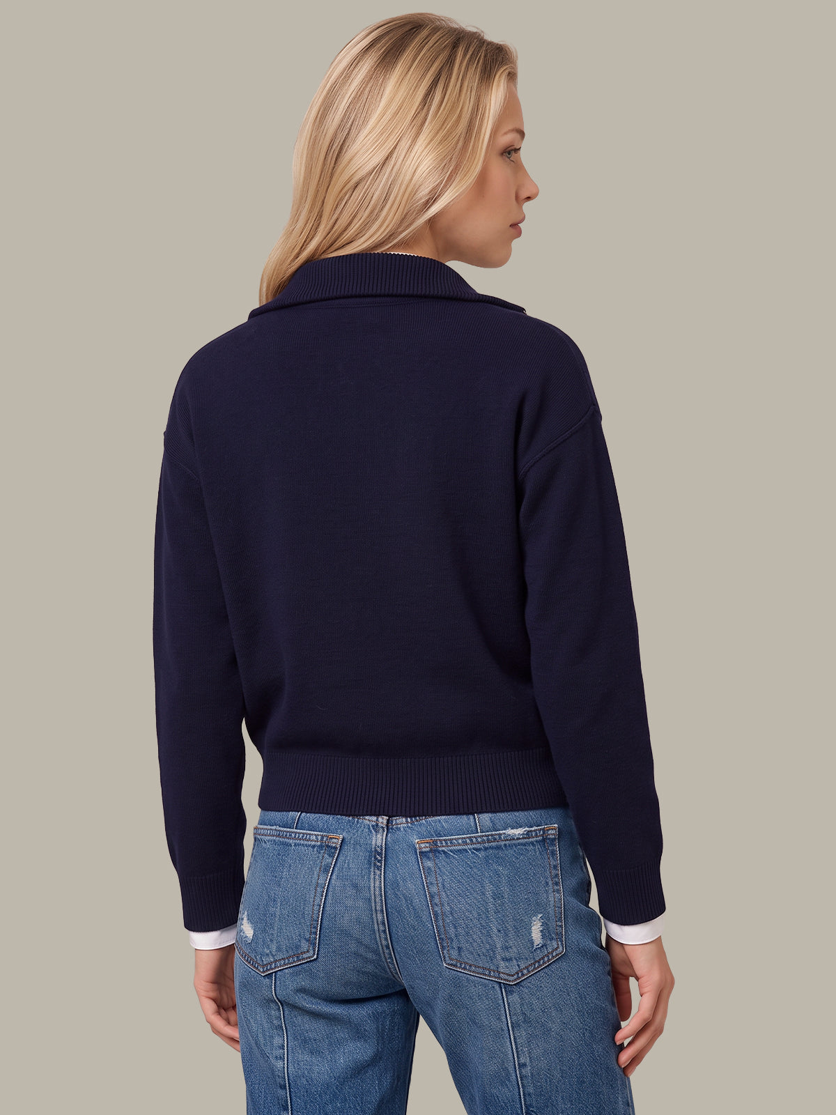 A woman shown from the back wearing a dark navy blue knit sweater with a collar and light blue jeans, set against a solid light gray background.