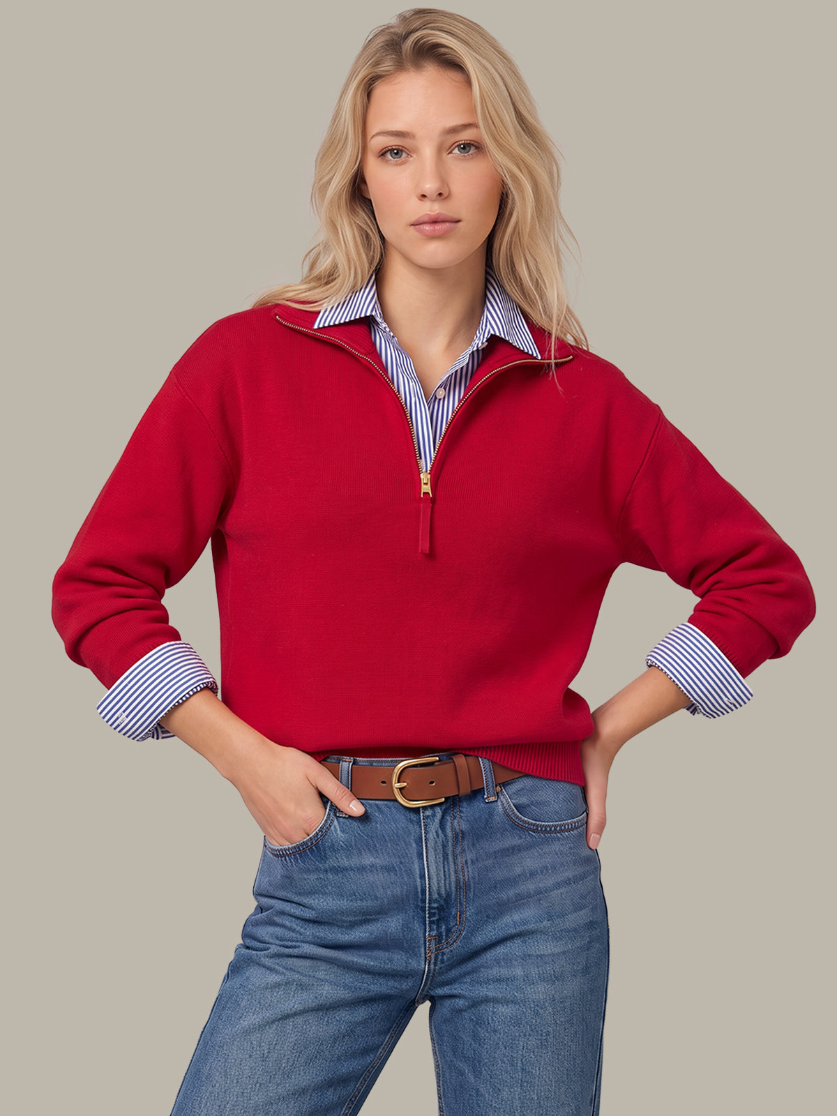 A woman modeling a red half-zip sweater layered over a blue and white striped collared shirt, paired with blue jeans and a brown belt, against a solid gray background.