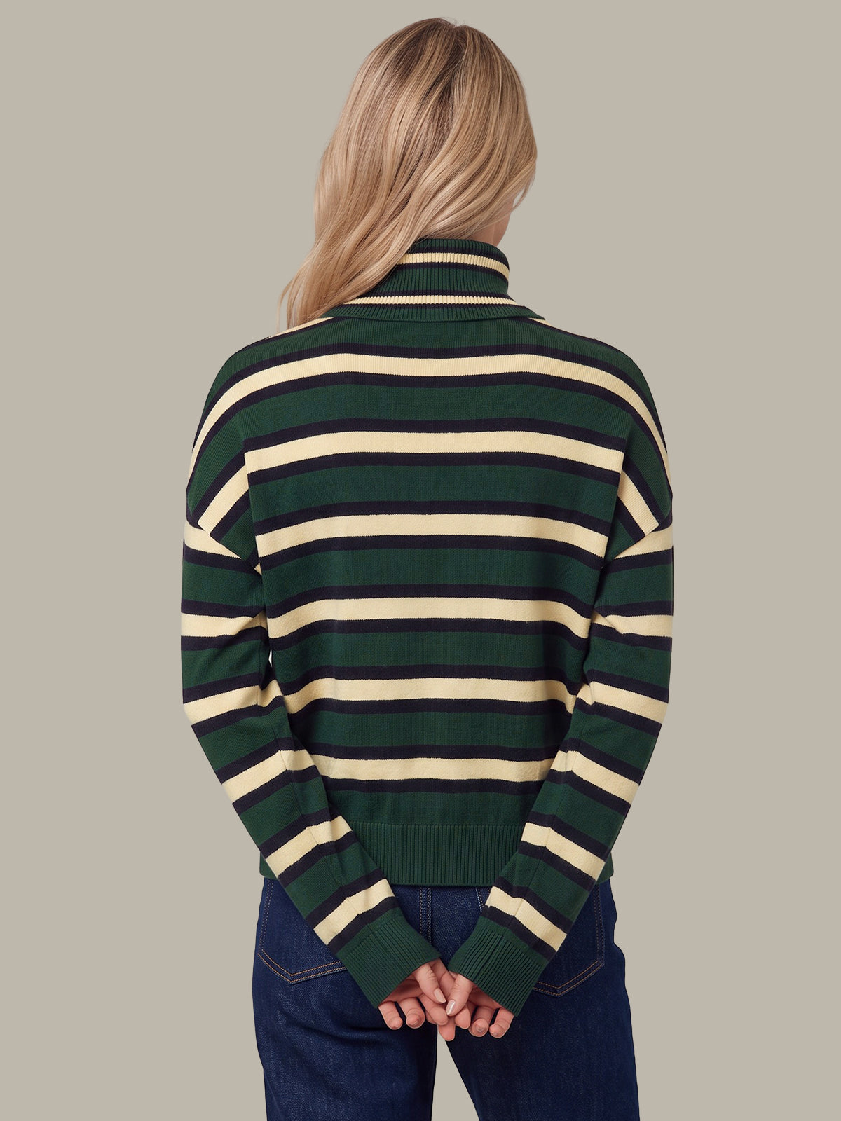 A woman viewed from behind wearing a dark green and cream striped turtleneck sweater and blue jeans, set against a solid light gray background.