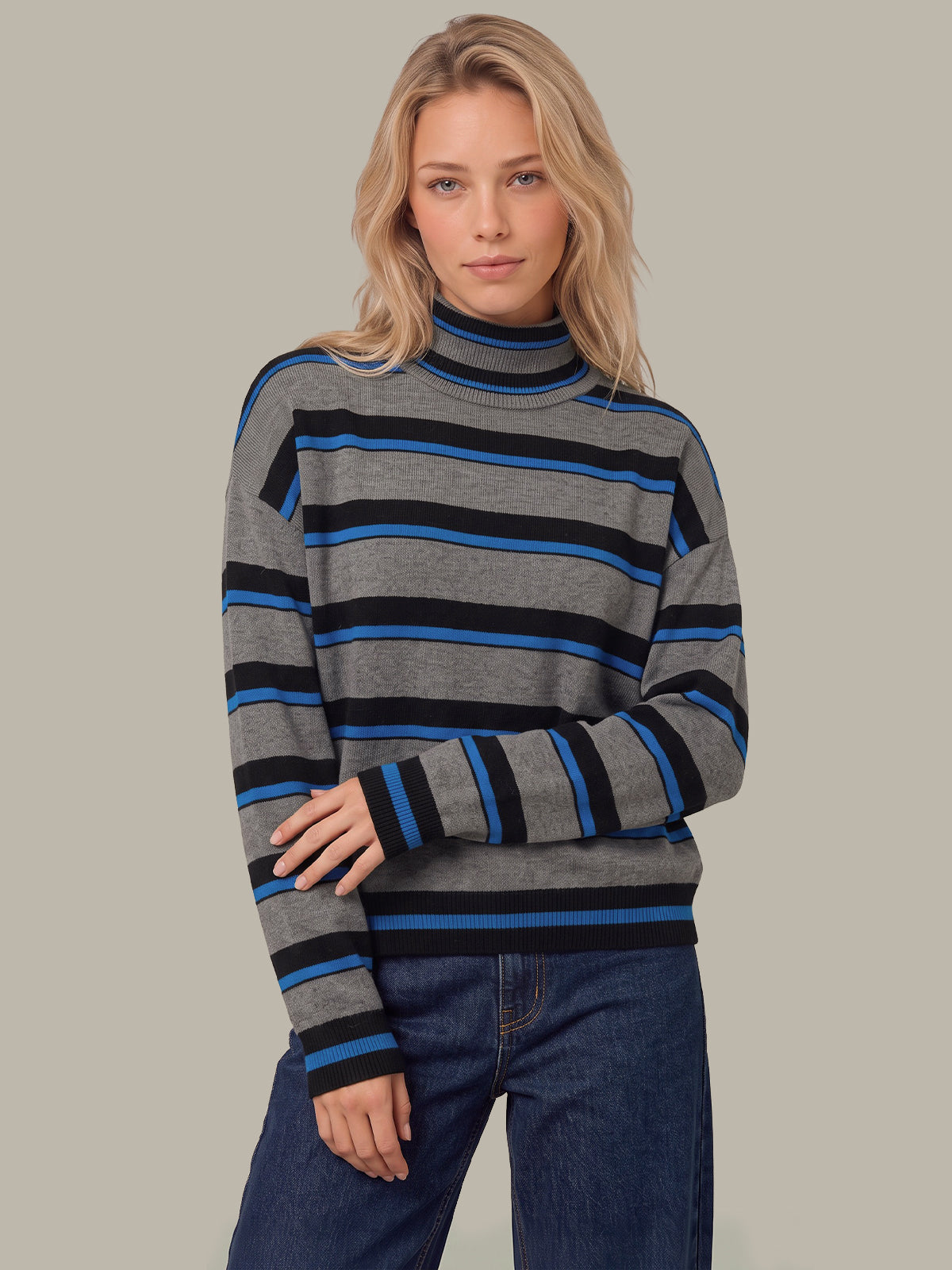 A blonde woman wearing a gray turtleneck sweater with horizontal black and blue stripes, paired with dark blue jeans, against a solid light gray background.