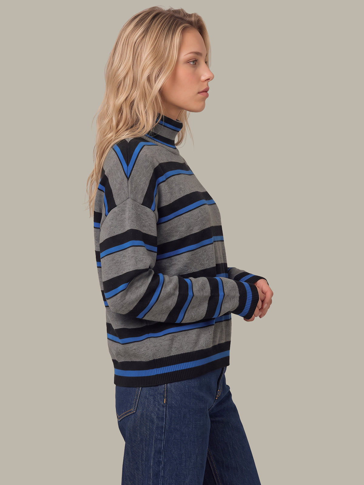 A woman wearing a gray knit mock neck sweater with horizontal black and blue stripes, paired with blue jeans, shown from a side profile against a solid light gray background.