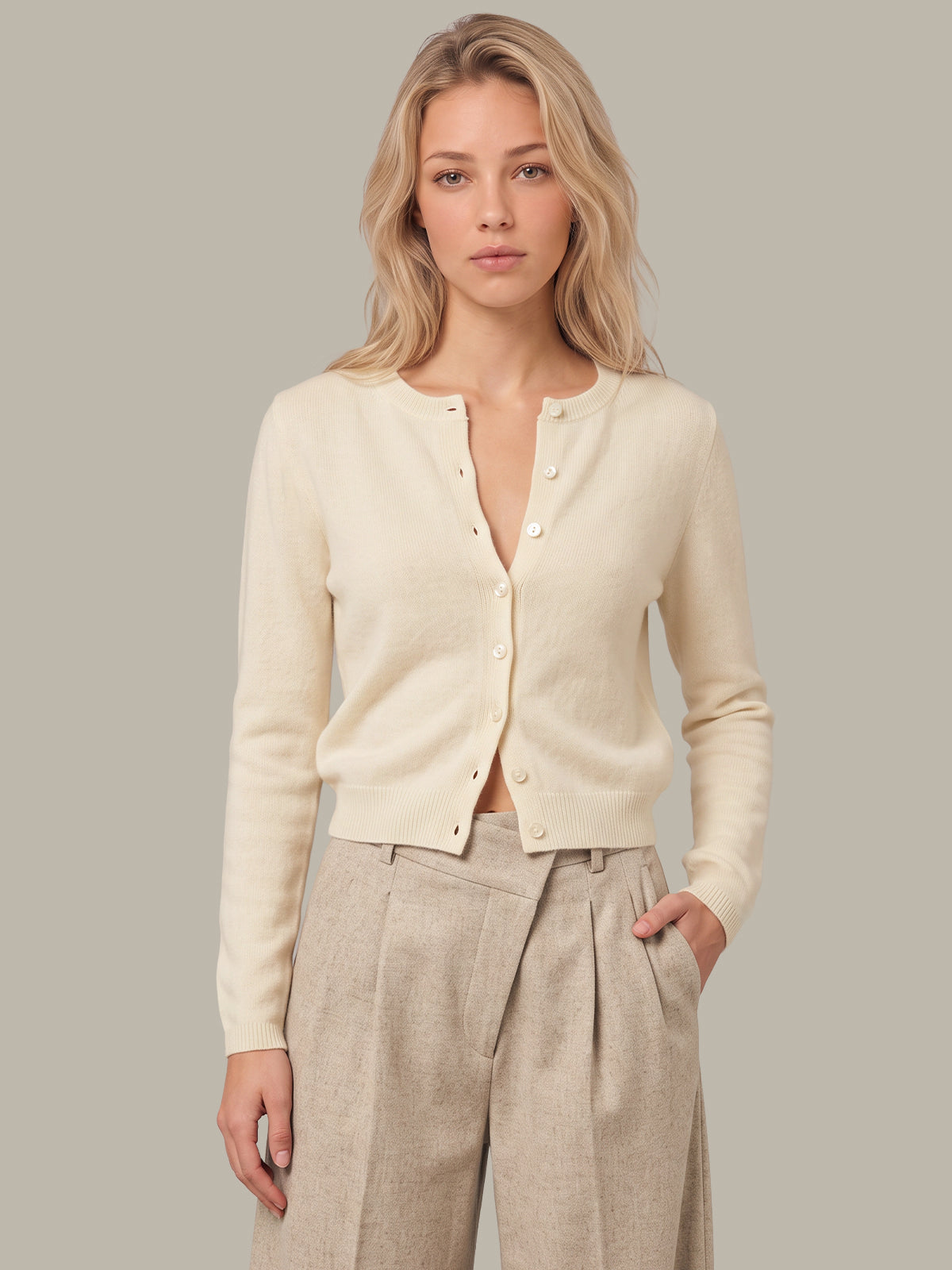 A blonde woman modeling a cream-colored, long-sleeve button-up cardigan sweater paired with light beige pleated trousers, set against a solid neutral gray background.