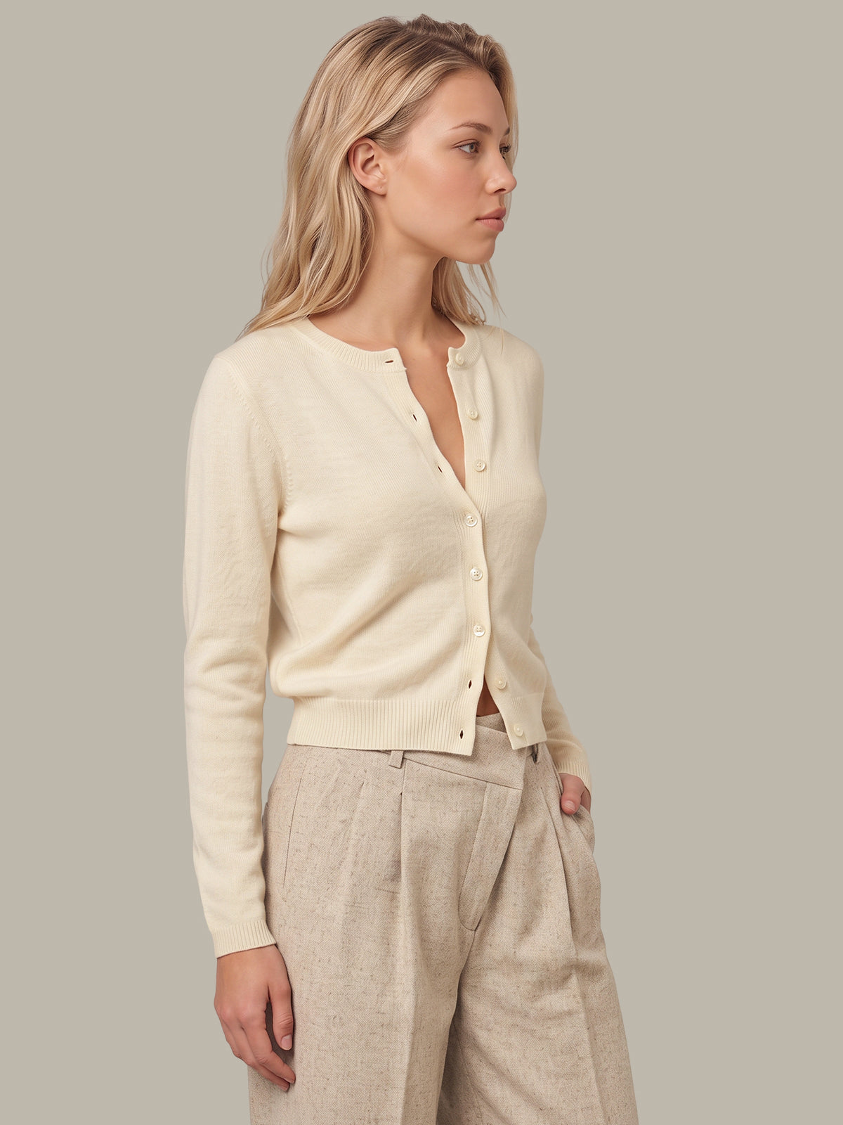 A woman wearing a cream-colored, long-sleeve button-up cardigan and light beige linen trousers, standing against a solid gray studio background.