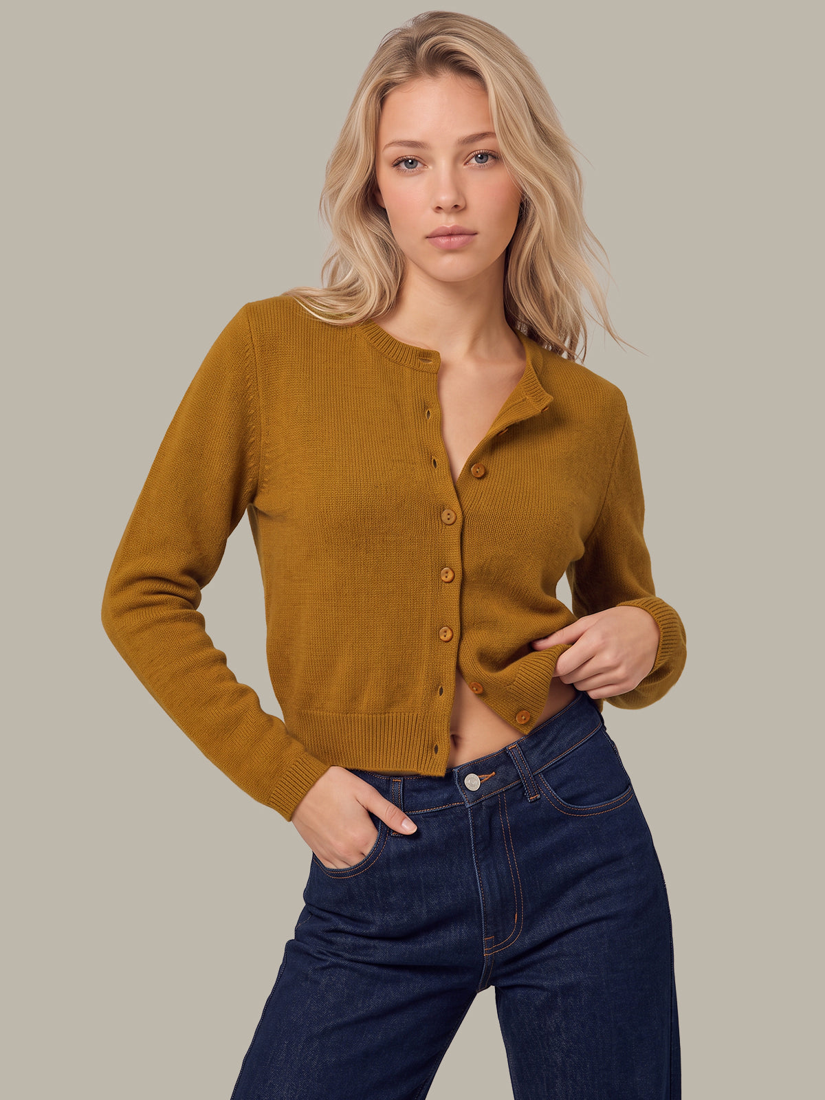 A blonde woman wearing a mustard yellow button-up cardigan sweater and dark blue jeans, posing against a solid light gray background.