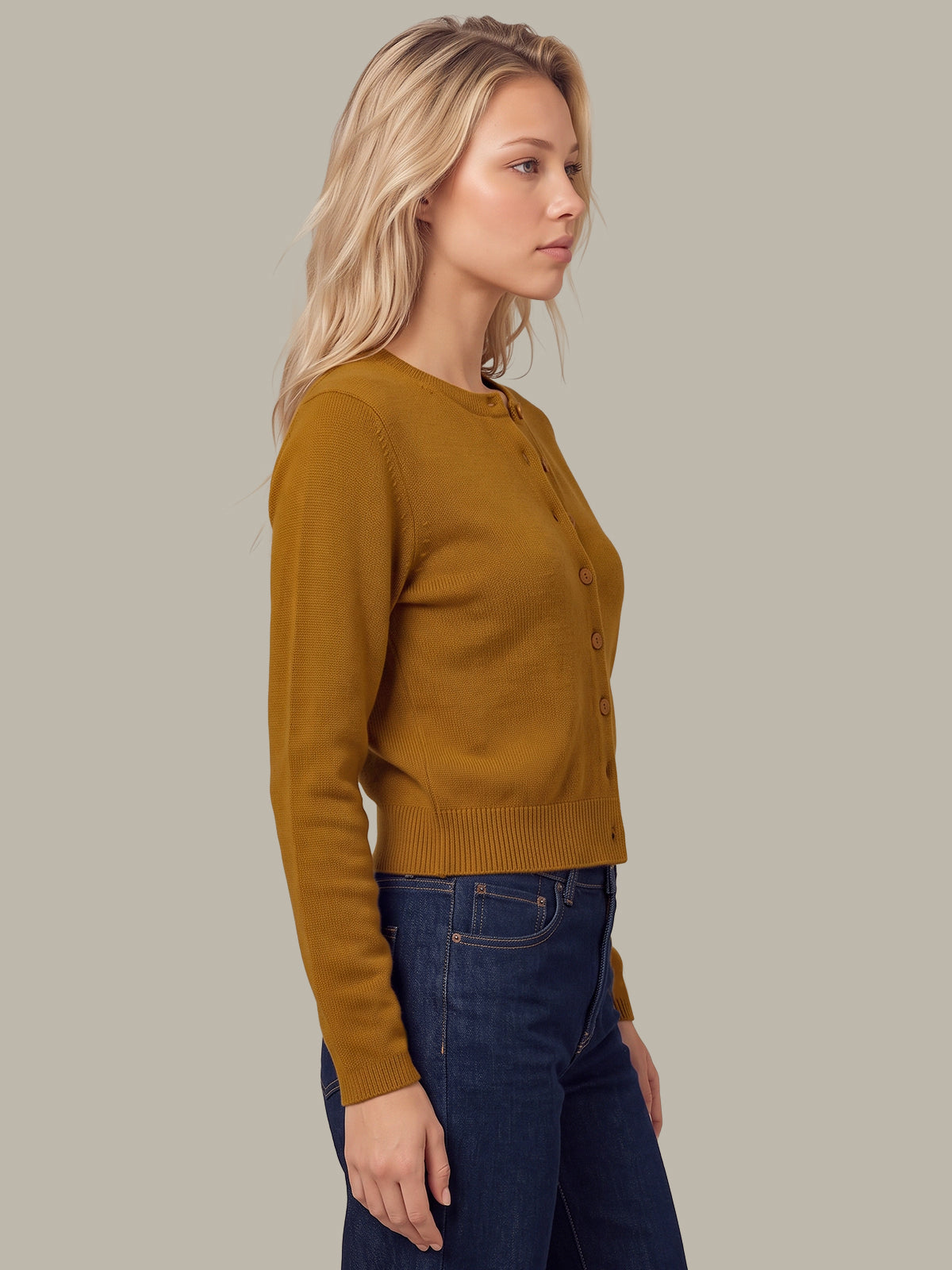 A woman in side profile wearing a mustard yellow knit cardigan with brown buttons and dark blue jeans, set against a solid light gray background.