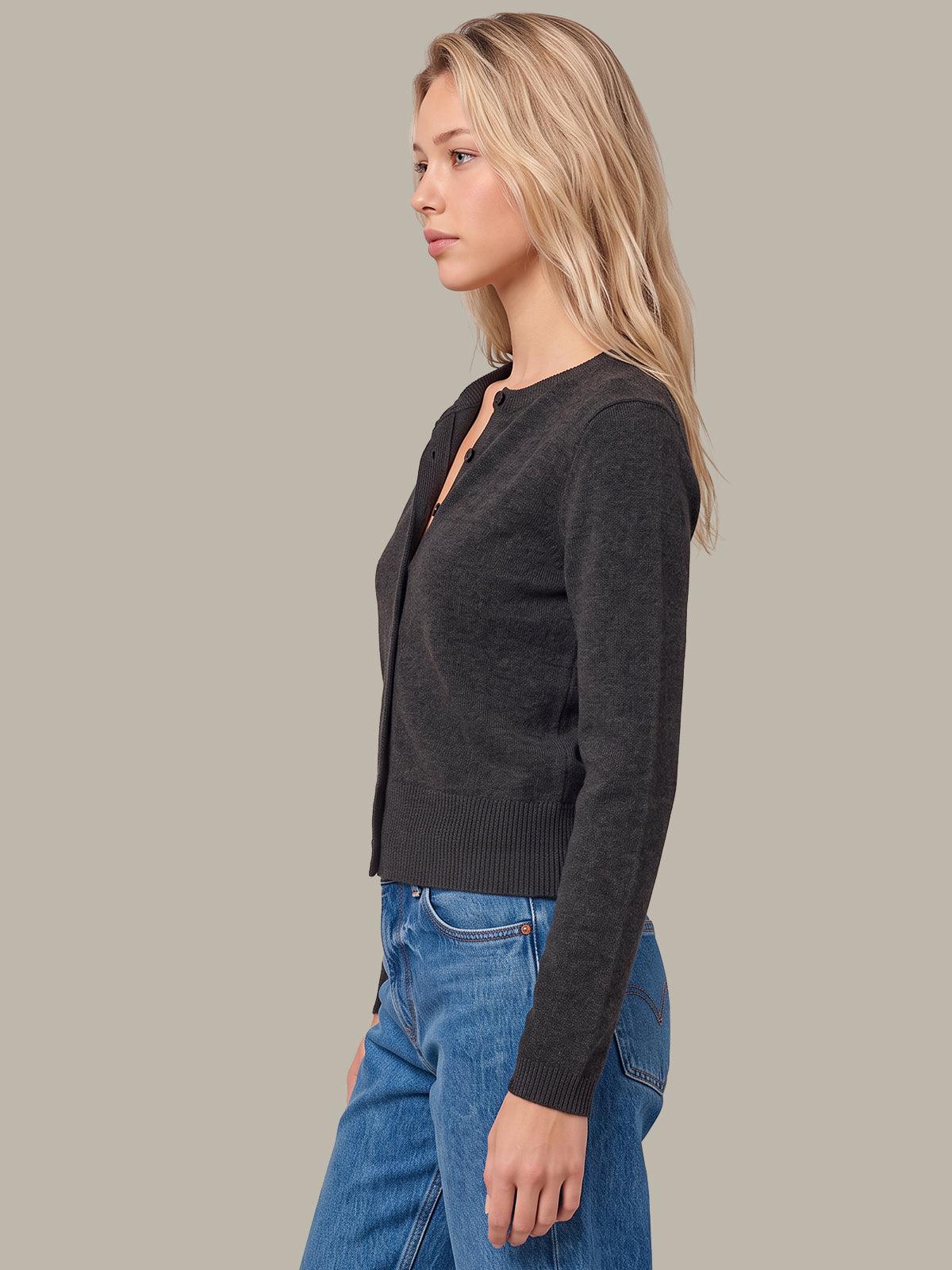 A woman in side profile wearing a dark charcoal gray long-sleeve knit cardigan with a split neckline over blue jeans, set against a solid light gray studio background.