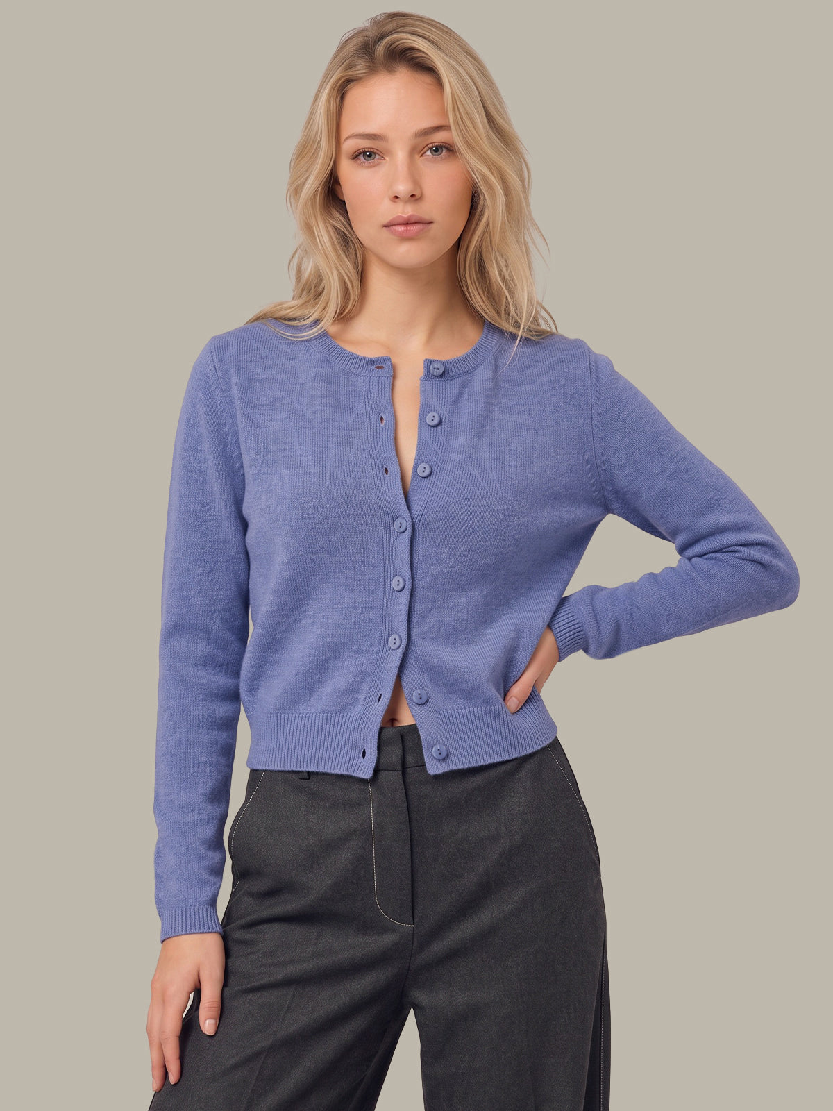A woman modeling a light periwinkle blue knit cardigan with a button-up front, paired with dark gray trousers, against a solid warm gray background.