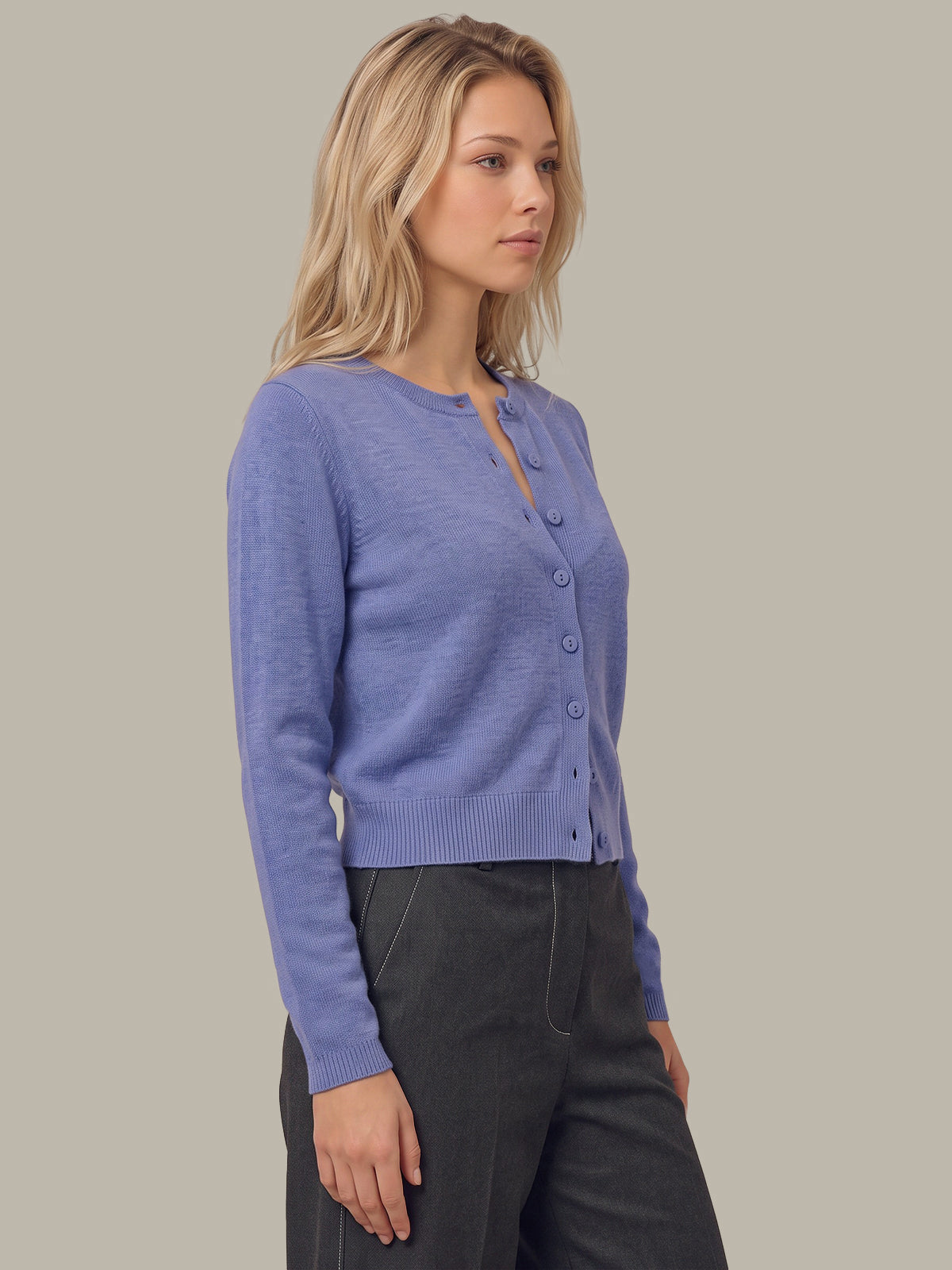 A woman wearing a light periwinkle blue button-up cardigan sweater and dark charcoal pants, standing against a solid warm gray background.