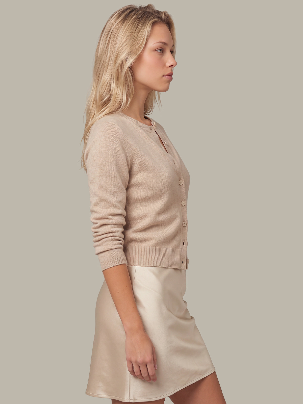 A woman in side profile wearing a light beige knit cardigan and a cream satin mini skirt, set against a solid light gray studio background.