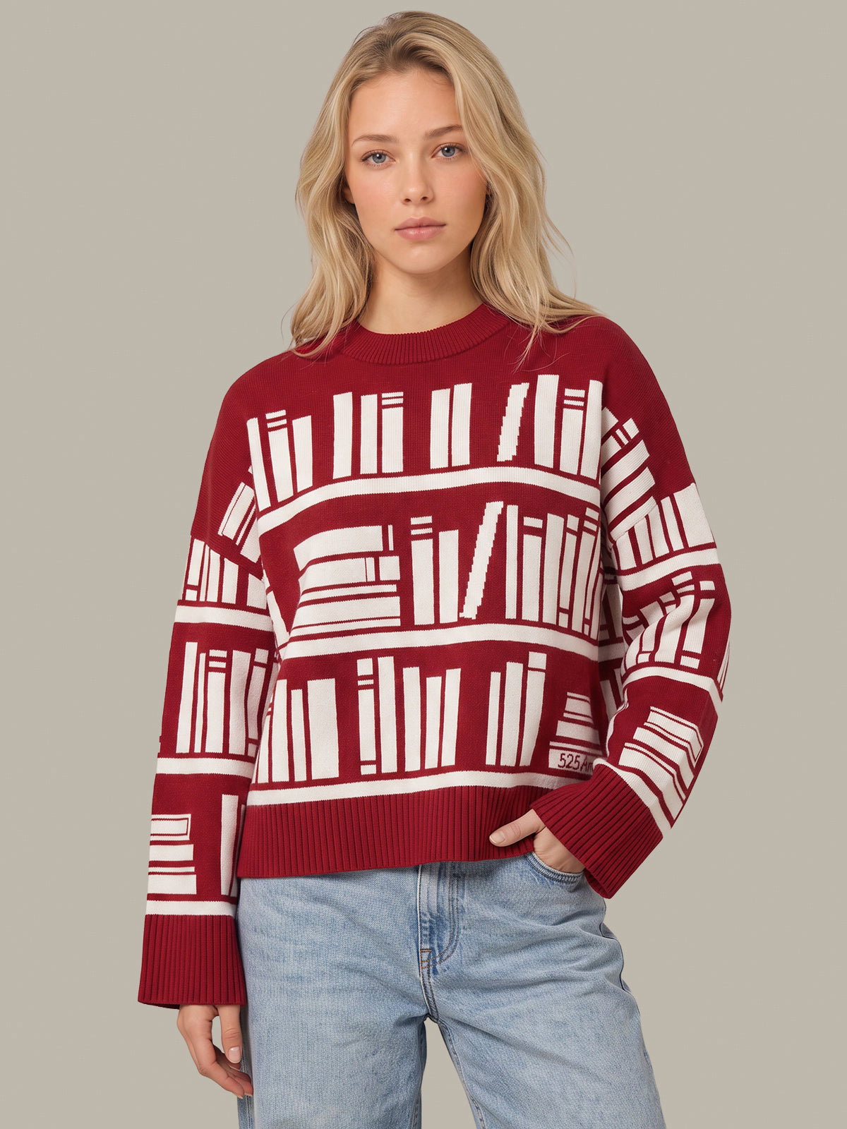 A woman modeling a red crewneck sweater with a white bookshelf and book pattern, paired with light blue jeans, against a solid gray background.